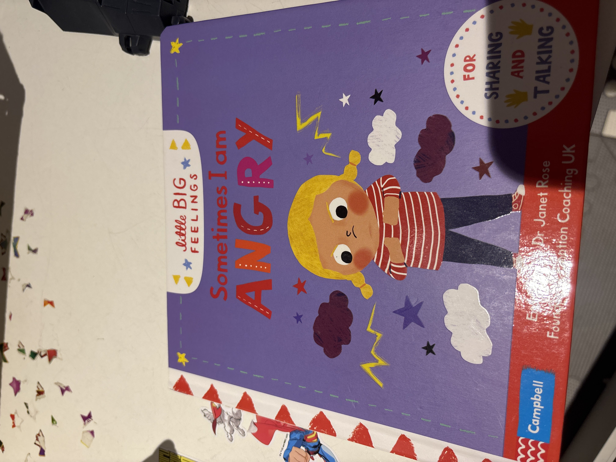 The image shows the cover of a children's book titled "Sometimes I am ANGRY" from the "Little Big Feelings" series. The book features an illustration of a young girl with blonde pigtails and a red and white striped shirt, with her arms crossed and a scowling expression. Around the girl are various drawings, including clouds, stars, and yellow lightning bolts. The book's cover is primarily purple with red and pink lettering. At the bottom of the cover, the publisher "Campbell" is visible in a blue rectangular logo, along with text indicating "Dr. Janet Rose" and "Foundational Coaching UK". A circular emblem on the lower right side of the cover reads "FOR SHARING AND TALKING". The book is placed on a surface with some scattered confetti-like shapes. The context of Dubai, United Arab Emirates, is not present in the image itself, as there are no identifiable landmarks, signs, or environmental cues to suggest this location.