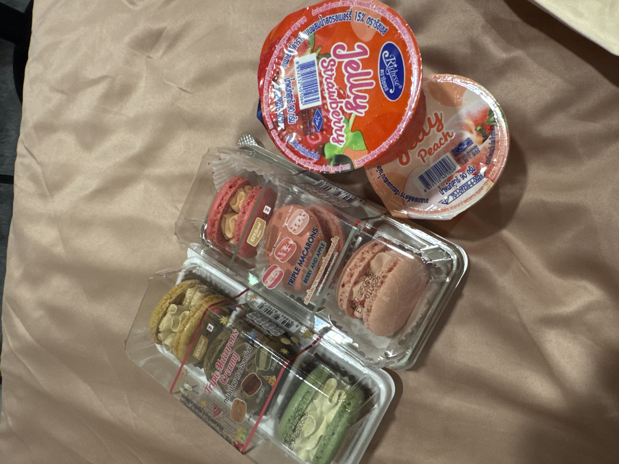 The image shows an arrangement of food items on a tan-colored fabric surface. There are two round containers of jelly desserts: one labeled "Jelly Strawberry" in orange packaging and the other labeled "Jelly Peach" in peach-colored packaging. Beneath the jellies, there are two rectangular plastic containers of "Triple Macarons". One container has macarons in red, brown, and pink shades. The other container has macarons in yellow, brown, and green shades. The packaging indicates these are "Creamy" and "Berry and Apple" flavors. The label suggests the macarons are made by "EZY Sweet" and the jelly desserts are "Richesse" brand. The text on the packaging is partly in Thai. The location is assumed to be somewhere in Kathu, Thailand, where these products are commonly sold.
