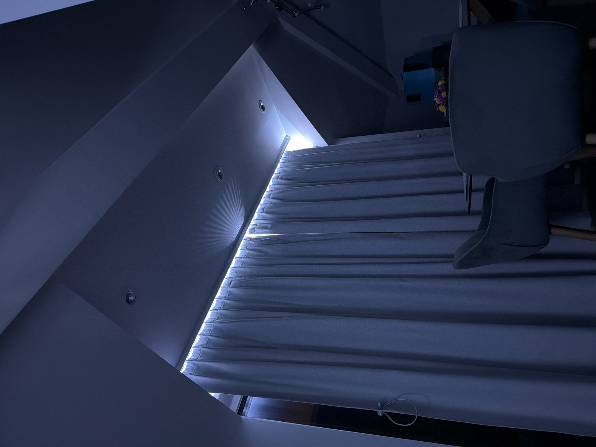 The image shows an interior space with a window covered by closed blinds, allowing light to penetrate through the slats. The walls and ceiling are painted in a dark blue color, with recessed lighting fixtures visible on the ceiling. Part of a chair is in the foreground on the right side of the image. The blinds and chair suggest the image was taken indoors. I have no information to suggest this image was taken in Buhovo, Bulgaria.