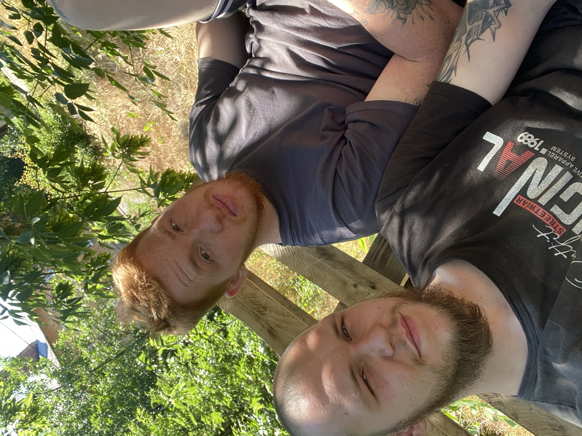 Two men are shown in a selfie outdoors. The man in the foreground is on the left and has a shaved head and a full beard. He is wearing a black t-shirt with white text. The man behind him is on the right, has red hair, and is wearing a grey t-shirt. He has tattoos on his arm. They are both seated on a wooden structure, possibly a bench, in a natural environment with green foliage and dry grass visible. The lighting suggests it is daytime, possibly summer, with sunlight filtering through the leaves. The background includes some greenery and what appears to be a building in the distance.