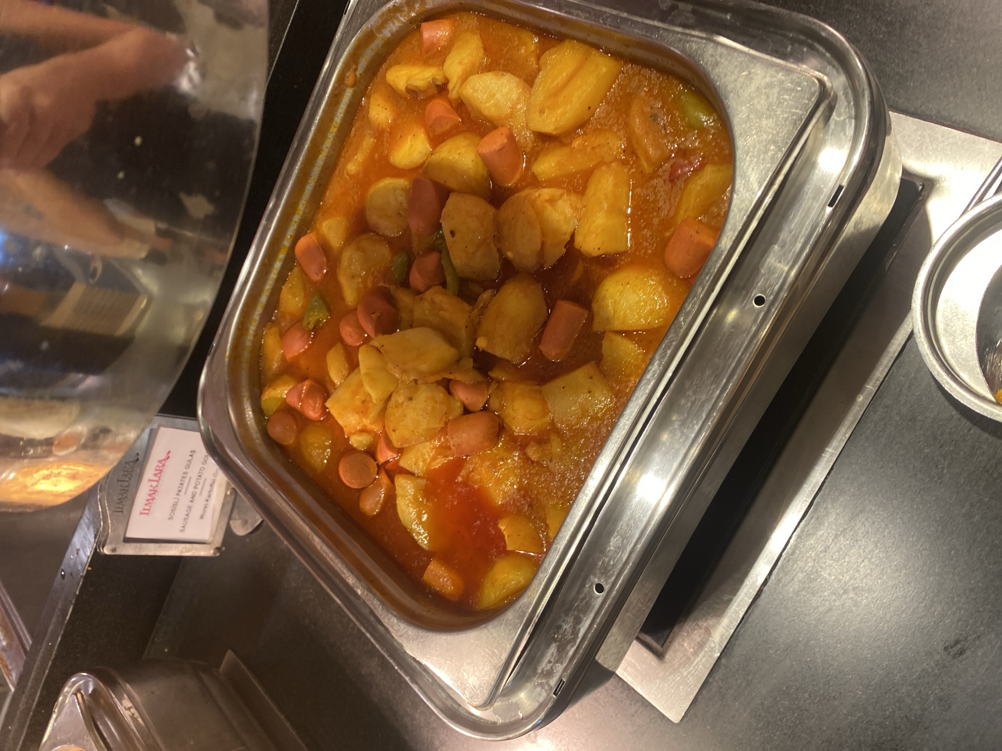 A close-up, eye-level shot shows a serving dish filled with a stew of chopped potatoes and sliced hot dogs in a red sauce. The dish is made of stainless steel and sits on a silver warming tray. A label is visible to the left of the dish with the text "ILMAK LARA" and below it "SOSISLI PATATES GULAS SAUSAGE AND POTATO GU". The background is dark and out of focus, with a hint of a reflective surface and a person's arm in the upper right corner. The lighting is bright, highlighting the food in the dish. The image depicts a buffet-style food service, possibly in a restaurant or catering establishment. The specific location in the city of Aksu, Turkey is not discernible from the image itself.