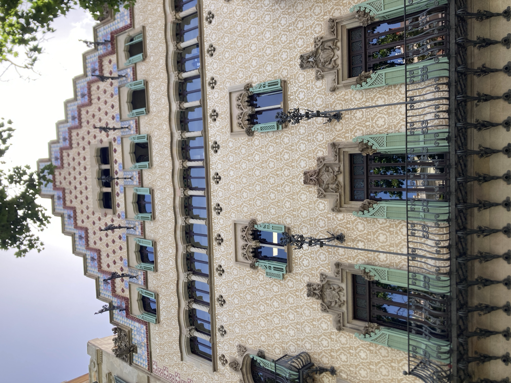 This is a vertical, eye-level shot of the facade of a building in Barcelona, Spain, possibly during daylight on a clear day. The architecture is ornate and detailed, featuring a stepped roofline on the left side adorned with a patterned mosaic of red, blue, and cream tiles. This pattern transitions into a repeating floral or geometric motif in cream on a beige background across the rest of the visible facade.
The building has numerous windows, many with dark frames and reflective glass that mirror the sky and surrounding trees. Some windows feature light green shutters with decorative grilles. Several balconies with intricate black wrought-iron railings are visible, with some extending outward from the facade. Decorative elements, such as sculpted figures resembling gargoyles and crests, are integrated into the stonework around the windows and cornices. The overall impression is one of rich historical or artistic architecture. No people are visible in the image, and no text is present.