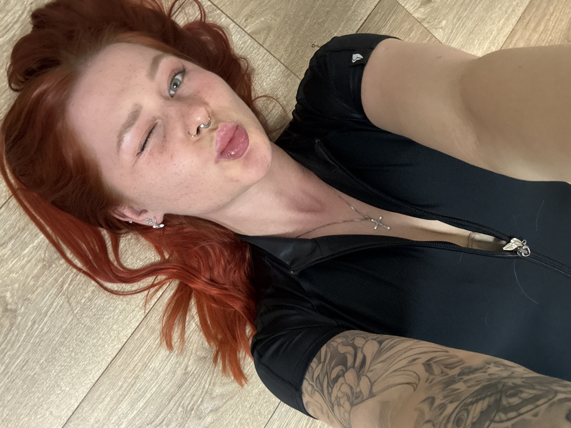 A woman with red hair is lying on a wooden floor. She is wearing a black zippered shirt. She is winking and making a kissing face. Her right arm, visible in the foreground, has a tattoo. A cross necklace is visible on her chest. There is no evidence to suggest that the image was taken in Rostov-on-Don, Russia.