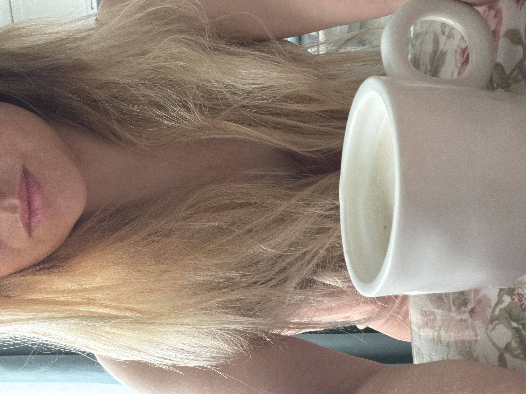 A close-up, eye-level shot presents a person with long, blonde hair draped over their shoulder, obscuring part of their chest. Only their lips and a portion of their shoulder are visible. A white mug filled with a light-colored liquid sits next to the person on a floral-patterned surface. The mug's handle is prominent. The image's background is out of focus. The location cannot be determined as Votkinsk, Russia from the provided image.