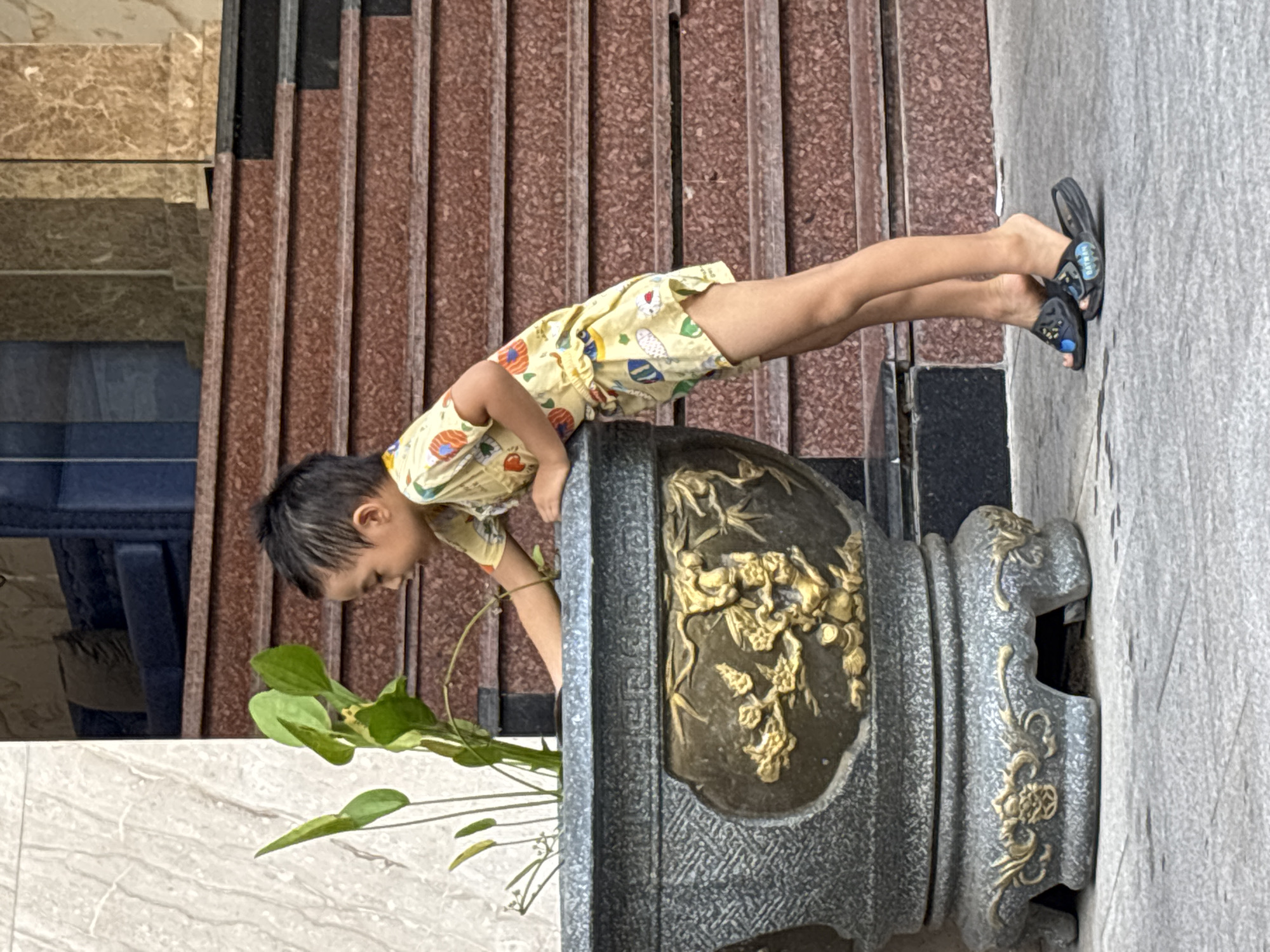 A young boy in a yellow shirt and shorts with cartoon prints, wearing black slide sandals, is reaching into a large, ornate stone planter with golden relief decorations. The planter contains a plant with green leaves. The boy is standing on a paved surface next to the planter. In the background, there are reddish-brown stone stairs leading up to a building with marble-clad columns. The scene appears to be outdoors in a city. The style of the planter and the marble cladding suggest a modern building with decorative elements, possibly in a public or residential area. The overall impression is one of a casual outdoor scene in an urban environment. The image does not contain any specific visual cues to pinpoint the exact location within Nha Trang, Vietnam.