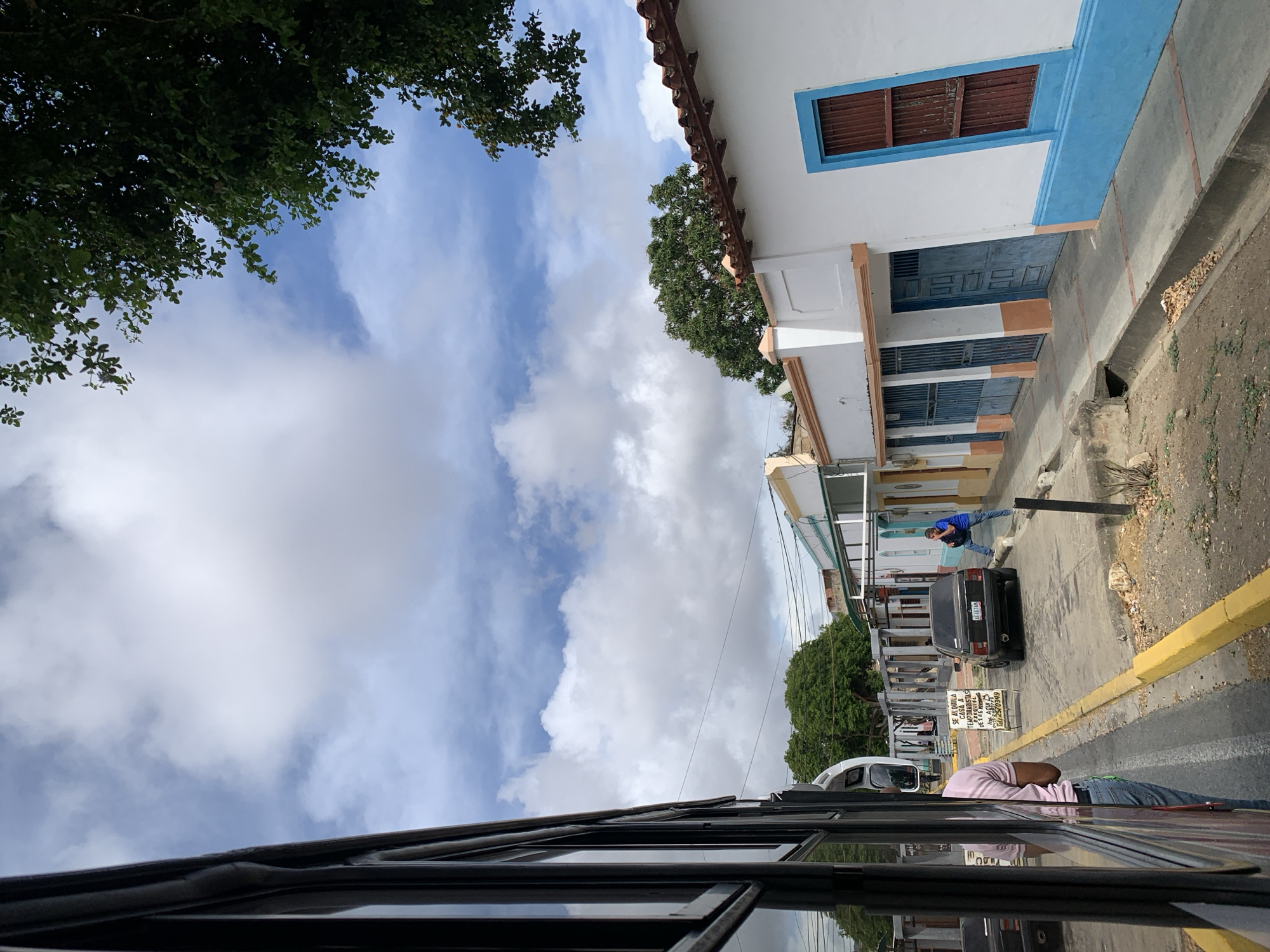 The image depicts a street scene in Tacarigua, Venezuela, captured from within a vehicle, likely a bus, as evidenced by the window frame in the foreground. A street lined with buildings is visible, painted white with blue trim and brown details. One man stands on the street talking on a cell phone. A black car is parked on the side of the road. A sign advertises rental services. The sky is cloudy with trees framing the top corners of the image.