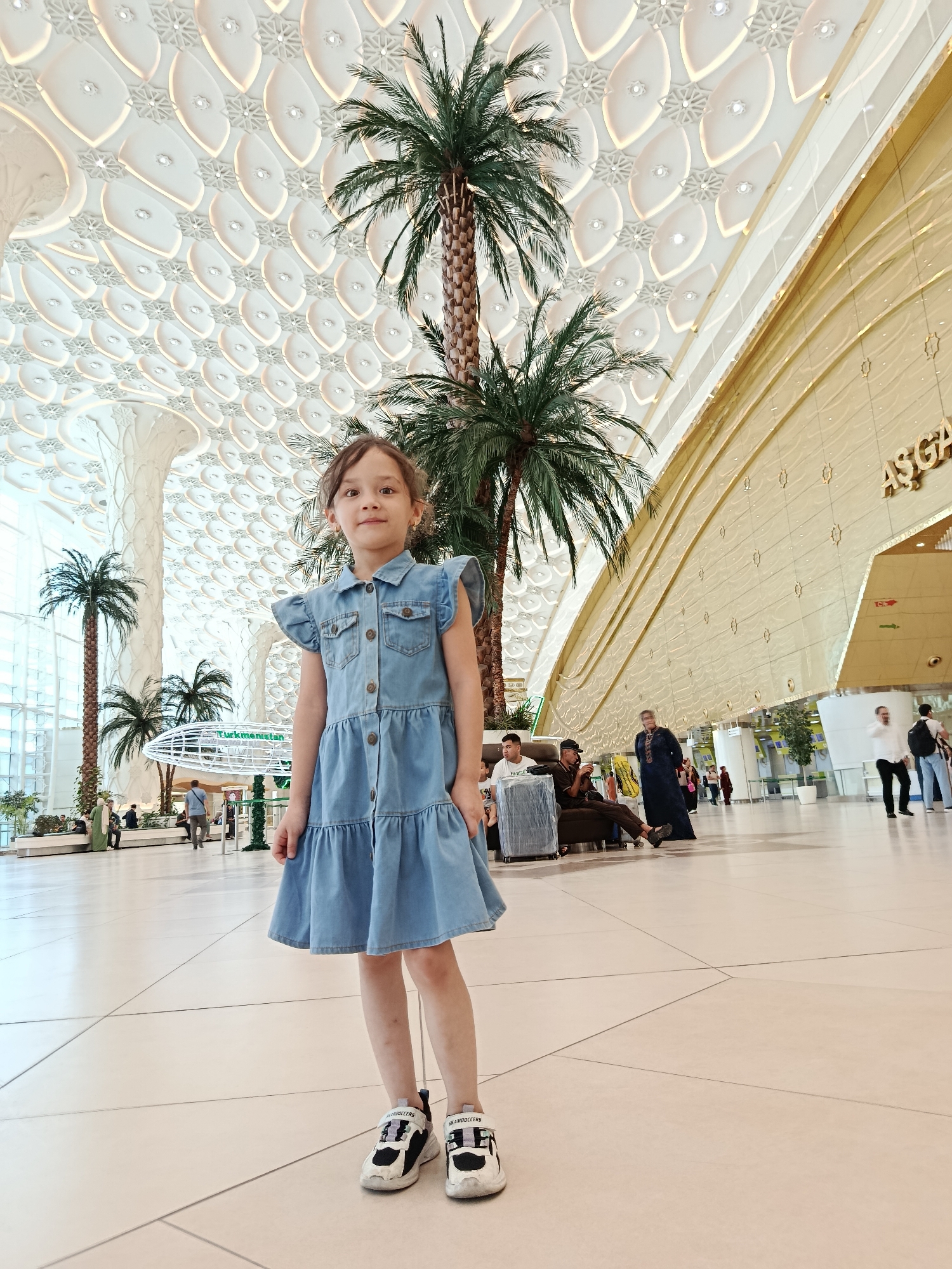 The image shows a young girl standing in a large, modern indoor space, likely an airport terminal in Ashgabat, Turkmenistan. She is wearing a denim dress and sneakers. Palm trees are visible in the background, along with architectural elements of the building. Other people are seen in the distance, some sitting and others walking. The ceiling has a distinctive patterned design. The overall impression is of a bright and spacious public area.