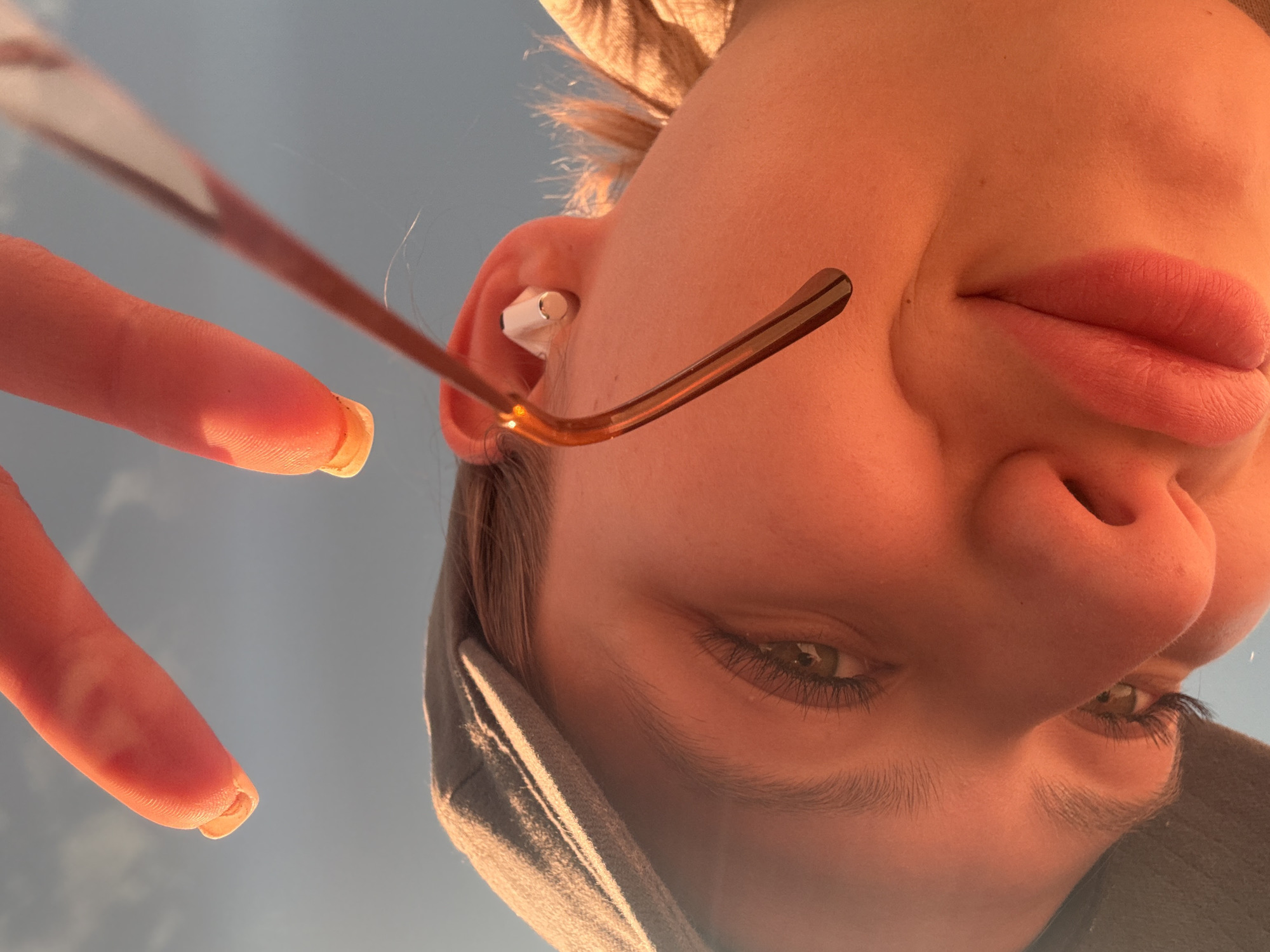 The image presents a close-up, low-angle perspective of a person looking directly downwards, likely at the camera. The person's face is illuminated by warm, golden light, indicative of either sunrise or sunset, casting an orange hue on their skin and hair.
The main subject is a young individual with green/hazel eyes, long eyelashes, and defined eyebrows. Their lips are full and have a natural reddish tint. They are wearing glasses with a thin, golden-brown frame, visible over the nose and extending towards the right ear. A white earbud is inserted into their right ear. Light brown or blonde hair catches the golden light around their face. A portion of a dark-colored or grey hoodie, possibly with textured or striped lining, frames the left side of their face.
In the upper left quadrant, a hand with manicured fingernails is visible, possibly holding the camera or gesturing. The fingernails are square-shaped with a light or natural polish, showing minor detail underneath. The background is a clear, light blue sky, suggesting fair weather. No text is visible in the image. The location context provided for this image is Bečići, Montenegro.