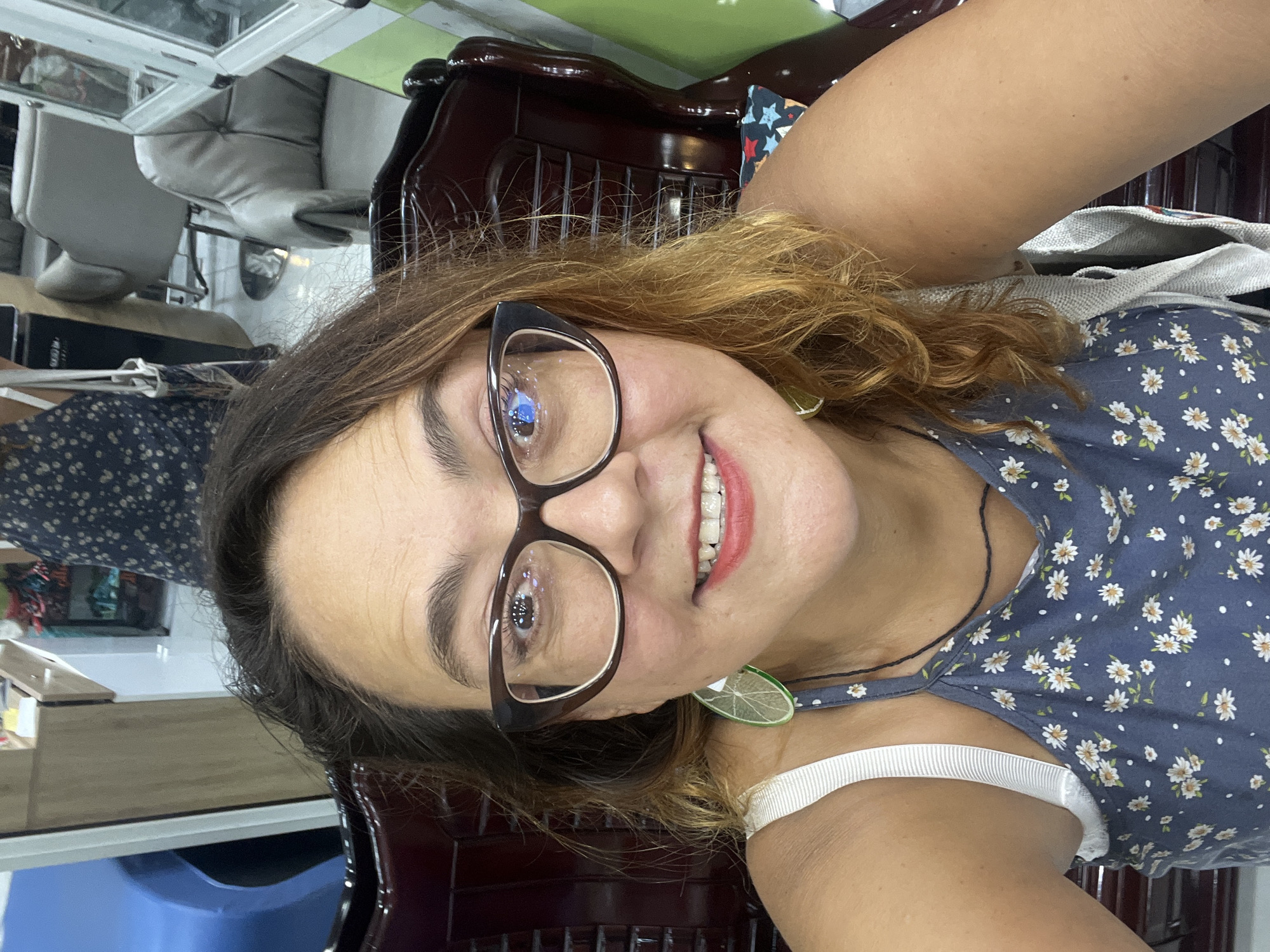 A medium shot selfie shows a smiling woman with medium-length auburn hair and glasses. She is wearing a blue floral-print dress with spaghetti straps. Her left earring is a lime slice. She is seated in a dark wood chair in what appears to be a salon or beauty parlor. In the background, there are chairs, mirrors, and shelves with products. The lighting is bright and natural. The location is Nha Trang Ward, Vietnam.