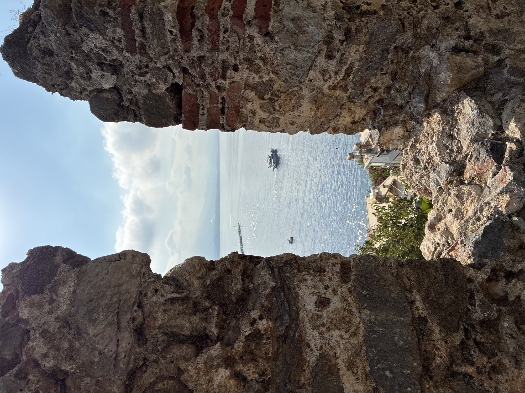 This image depicts a view through an opening in ancient stone ruins in Alanya, Turkey. The opening frames a sunlit seascape with a bright blue sky and scattered clouds. A pleasure boat is visible on the water, moving away from the viewer. In the distance, a pier extends into the sea, and the faint outline of the coastline and mountains can be seen. Below the waterline, part of a town with buildings and trees is visible, suggesting the ruins are situated on a hill overlooking the city. The stone walls in the foreground are weathered and show signs of age, with visible layers of different materials, including some red brickwork. The sunlight creates a sparkling effect on the surface of the water.