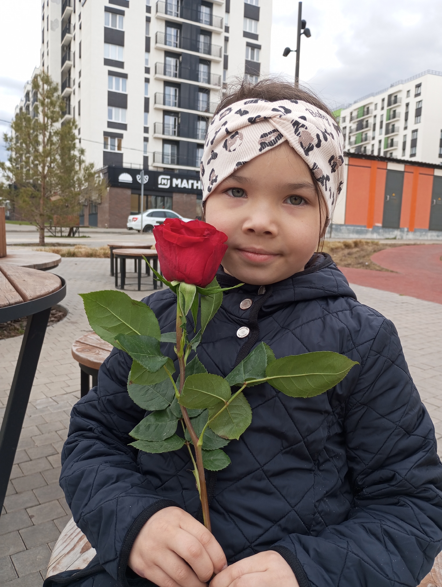 A young girl with light-colored eyes and dark hair, wearing a cream-colored headband with a leopard print pattern and a dark blue quilted jacket, is seated outdoors, looking directly at the camera. She holds a single long-stemmed red rose with green leaves in both hands, positioned centrally in the frame. Her expression is gentle and includes a slight smile.
The scene takes place in an urban or suburban environment, likely within a modern residential complex in Pervomaysky, Russia. In the background, several tall, multi-story residential buildings with predominantly white facades and dark accents (balconies, window frames) are visible under an overcast sky. A green, coniferous tree stands to the left of the frame.
The foreground shows paved ground and outdoor seating, including a round wooden table with matching stools. Further back, a white car is partially visible on a street, alongside a commercial building with a dark facade. On this building's sign, partial text "МАГН" is clearly visible, suggesting it is a "МАГНИТ" (Magnit) store. Faint, stylized text resembling "Baby" can also be observed on the girl's headband. The overall impression is a daytime scene, possibly on a cool day given the girl's jacket.