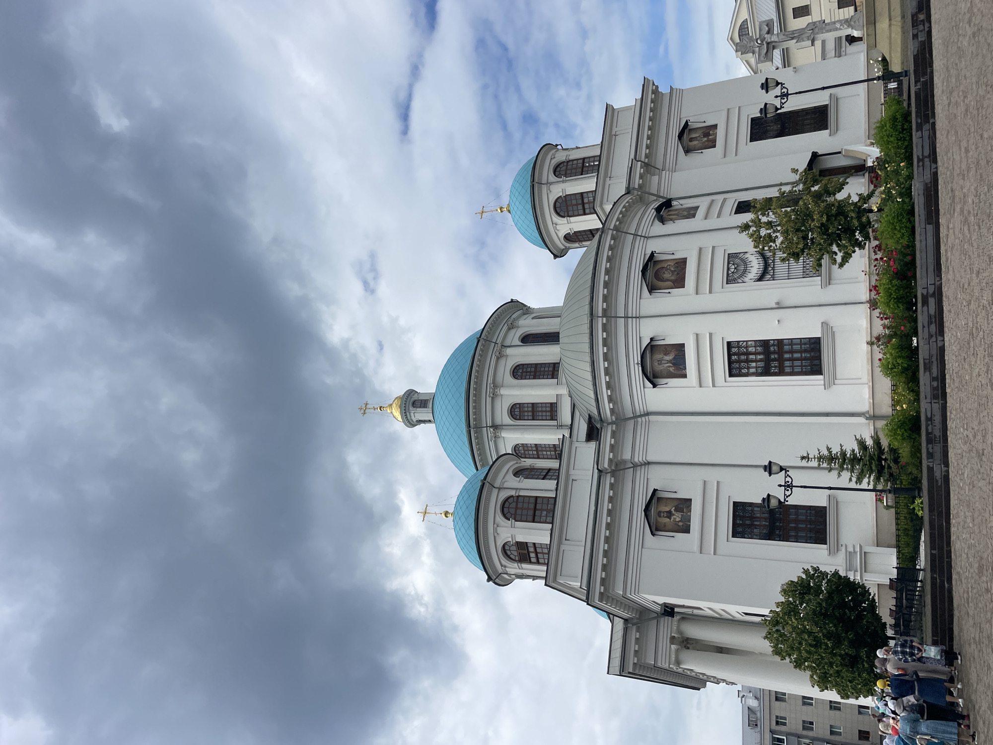 This is a low-angle, eye-level, exterior, full shot of the Kazan Kremlin's Cathedral of the Annunciation on a cloudy day. The white, neoclassical structure features several bright turquoise domes topped with golden crosses. Icons are displayed in framed niches on the exterior walls. People are visible in the lower left corner, gathered on a cobblestone plaza in front of the cathedral. Green trees and flower beds are also present in the foreground. The sky is filled with dramatic, dark grey clouds interspersed with patches of brighter blue sky.