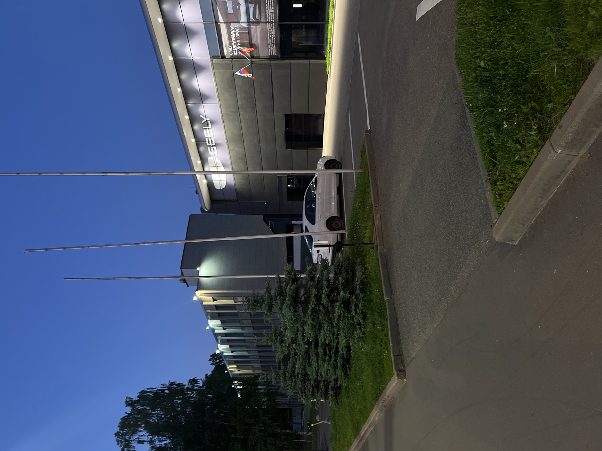 This forensic image captures an exterior view of a commercial area in Saint Petersburg, Russia, at dusk or night, indicated by the deep blue sky and illuminated artificial lighting.
The dominant structure on the right is a modern Geely car dealership, identifiable by the large "GEELY" logo displayed prominently on its dark grey facade. A large glass window on the right section showcases a vehicle, with Cyrillic text visible, including "GEELY COOLRAY" and "ЭНЕРГИЯ ГОРОДА" (Energy of the city). A Russian flag is affixed vertically to the building near this window. Below the main facade, two large, dark garage doors are visible. A white sedan is parked in a marked space directly in front of the dealership.
To the left of the dealership stands a multi-story building with a more classical or industrial design, featuring vertical architectural elements illuminated by warm-toned lights. In the mid-ground, a row of well-maintained evergreen trees (likely spruces) is planted in a grass verge, separating the two buildings. Three tall, slender metallic poles, presumably flagpoles, rise from this grass verge, devoid of flags. Further left, beyond the trees, a yellow and black electric scooter is parked on the grass, and a faint green light is visible.
The foreground consists of asphalt pavement, including a parking area delineated by white lines, and concrete curbs separating the pavement from patches of green grass. No people are present in the scene.