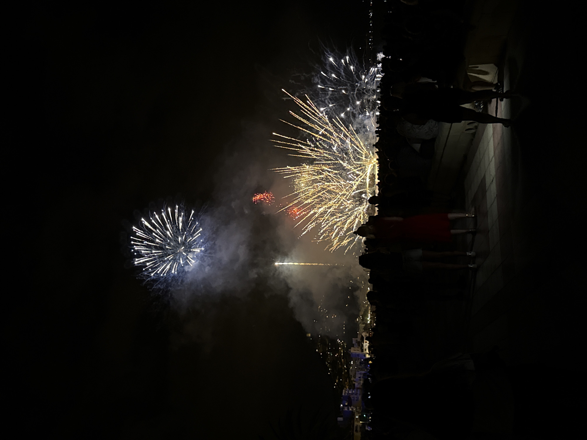 This is a vertical, outdoor, low-angle shot of a fireworks display occurring at night over the city of Blanes, Spain. In the foreground, the silhouettes of a crowd of people are visible, facing the fireworks. They are standing on a paved area with a low wall to their right, where some individuals are seated or leaning. Further in the background, the city lights of Blanes are scattered across a hillside. The sky is filled with multiple bursts of fireworks, some white and some gold, with plumes of smoke obscuring parts of the display and the cityscape. One prominent firework in the upper left is white with a feathery, starburst pattern. To the right, a dense cluster of gold fireworks radiates outwards, with smaller white fireworks interspersed. A single, thin vertical streak of light, possibly a firework ascending, is visible above the city. The overall atmosphere is festive and dark, illuminated by the bright explosions of fireworks.
