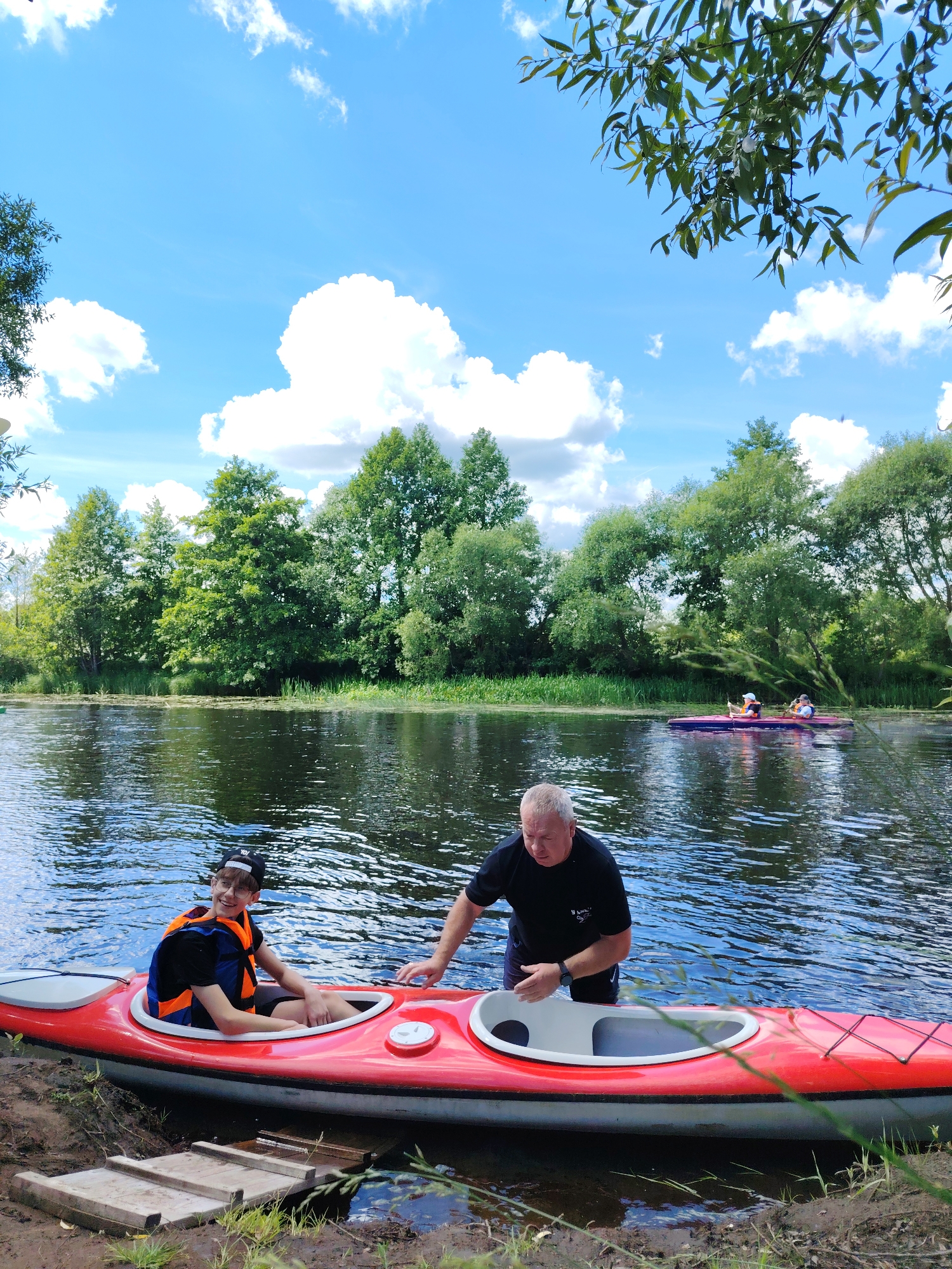 Two people are near a red kayak on the bank of a body of water, likely a river or lake, in Мётчанскі сельскі Савет, Belarus. A young person wearing glasses and an orange and blue life vest is seated in the front cockpit of the kayak. An adult male wearing a black t-shirt and dark shorts is standing next to the kayak, with his right hand reaching towards the rear cockpit. The water is calm with ripples, and the surrounding environment features lush green trees and vegetation along the shoreline under a bright blue sky with scattered white clouds. In the background, two more kayaks with people in them are visible on the water.