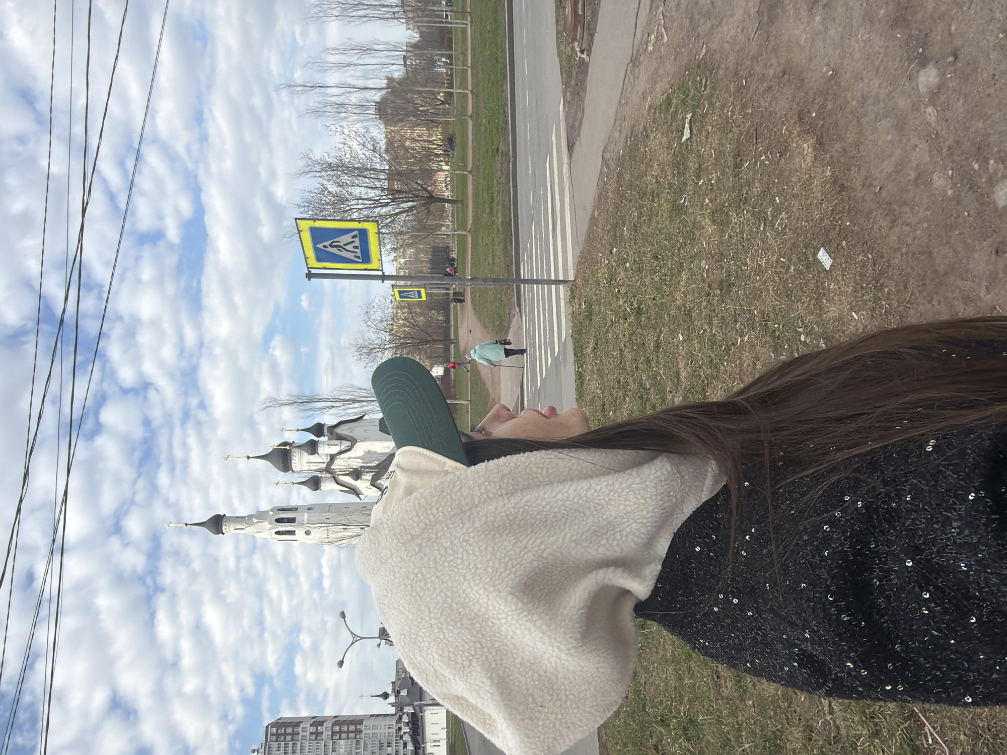 The image depicts an outdoor urban scene, featuring a person in the foreground looking upwards, and a street with a prominent Orthodox church in the background.
The main subject, a person with long dark hair, is seen in profile, wearing a dark green baseball cap, a cream-colored, fluffy-textured hoodie or jacket, and a black garment adorned with sequins on the visible shoulder and arm. Her gaze is directed towards the sky or the distant church.
In the midground, a road with a white-striped pedestrian crossing is visible. Another individual, wearing a light-colored jacket and using a walking stick, is crossing the street. Trees with bare branches, suggesting early spring or late autumn, line the roadside.
The background is dominated by a large, white Orthodox church featuring multiple black onion domes and a tall bell tower, indicative of Russian architectural style. Other buildings are faintly visible further in the distance. The sky above is partly cloudy with patches of blue.
Overhead power lines crisscross the upper left of the frame. Two yellow-and-blue pedestrian crossing signs, depicting a walking figure, are clearly visible. The ground in the foreground shows patches of dry grass and dirt, with some scattered debris. Given the prominent Orthodox architecture, the location aligns with the provided context of Saint Petersburg, Russia.
