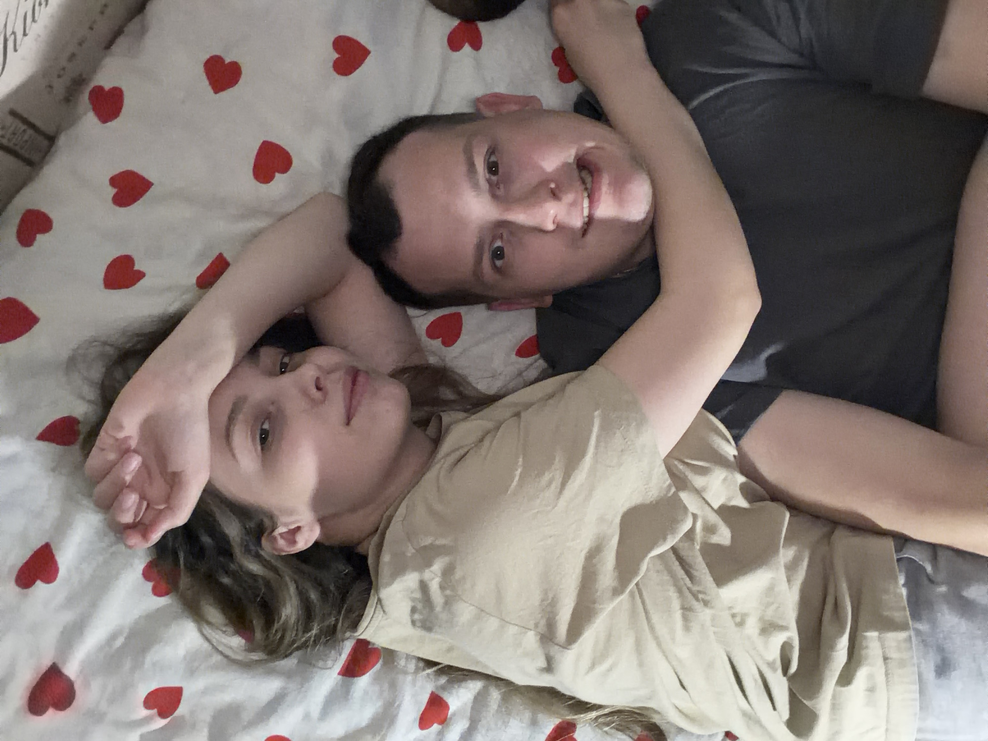 Two people, a man and a woman, are lying on a bed with a white sheet covered in red heart patterns. The man is wearing a dark gray t-shirt and is smiling. The woman is wearing a tan t-shirt. Both have an arm raised above their heads. The location is Chaikovsky, Russia.