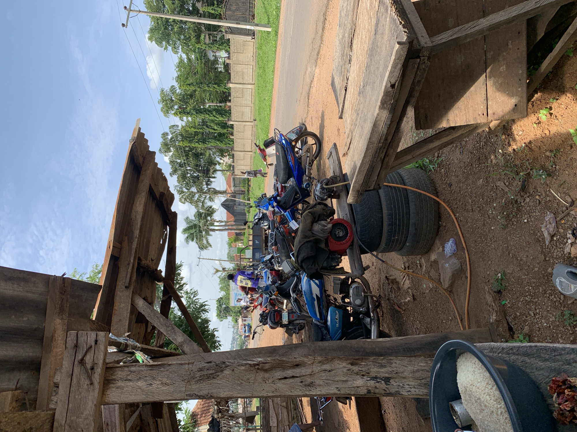 The photograph depicts a scene at a roadside stall in Gbeleko, Nigeria, on a bright, partly cloudy day. In the foreground, a large, rough-hewn wooden post supports a corrugated metal roof structure. Beneath the shelter, several motorcycles are parked on the dirt ground. A blue motorcycle with a black seat is prominently featured, with a worn black jacket draped over its rear. To the left, a dark basin filled with white rice sits on a wooden surface, next to some dried red objects. Stacked tires are visible behind the basin.
Beyond the immediate stall area, a paved road runs horizontally across the image. A few individuals are present on the road and sidewalk. One person in a bright purple jacket stands near a truck on the right side of the road, while another figure in gray walks further down the road on the left. A long concrete wall with a decorative iron gate runs along the right side of the road, bordered by green grass and tall trees, including palm trees. Power lines are visible against the sky. The overall impression is one of a busy, informal commercial area situated alongside a main thoroughfare in a Nigerian town.