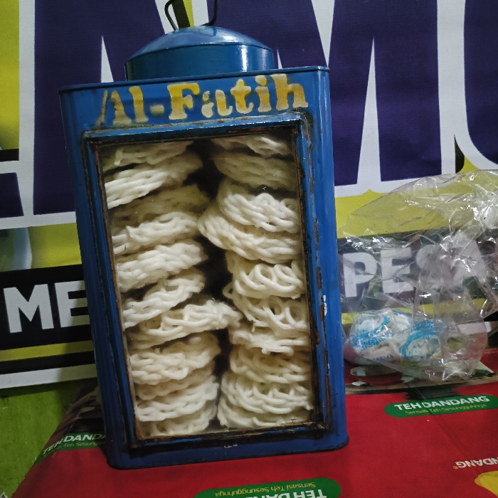 The image shows a blue metal box with a glass window containing a stack of white, circular, crispy crackers. The box is labeled "Al-Fatih" in yellow lettering. The box is sitting on a red and white surface with a plastic bag containing white items. The coordinates (latitude: -6.975, longitude: 110.332) indicate that the location is in Tugu, Indonesia. The surface behind the box is a blue and yellow wall with writing in Indonesian.