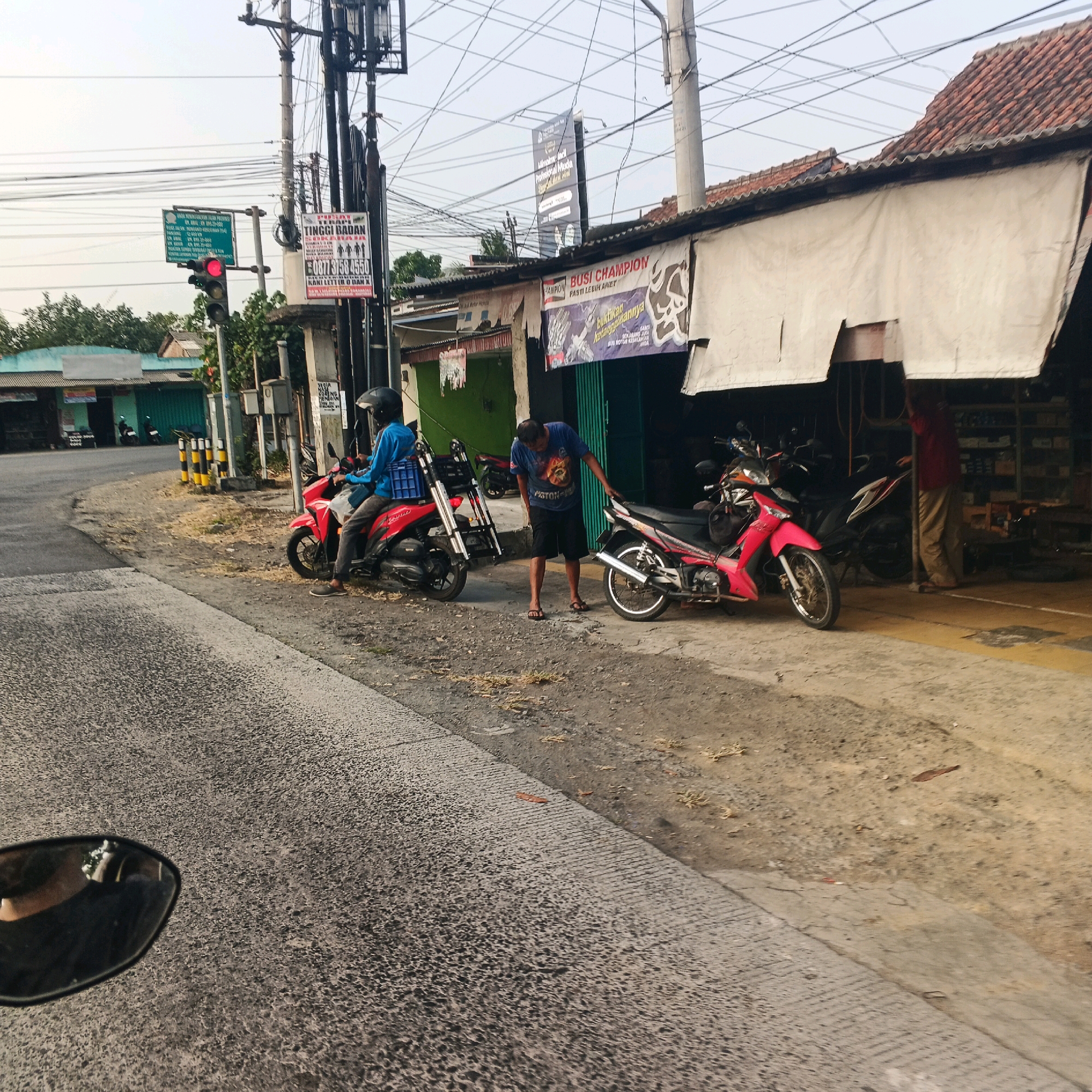 The image depicts a street scene in Menganti, Indonesia, likely near a commercial area, with a traffic light showing red. In the foreground, a person wearing a blue shirt and a black helmet is seated on a red motorcycle, facing away from the camera. The motorcycle has a blue crate attached to its rear. To the right of this motorcycle, another person in a blue t-shirt and black shorts is standing and leaning towards a pink motorcycle. This second motorcycle is parked under an awning in front of a building that appears to be a shop, indicated by shelves stocked with items visible through an open doorway. The shop's facade is decorated with banners. A power pole with numerous wires stands prominently in the background. The ground is paved with asphalt and dirt. The time of day appears to be late afternoon or early evening, with soft lighting.