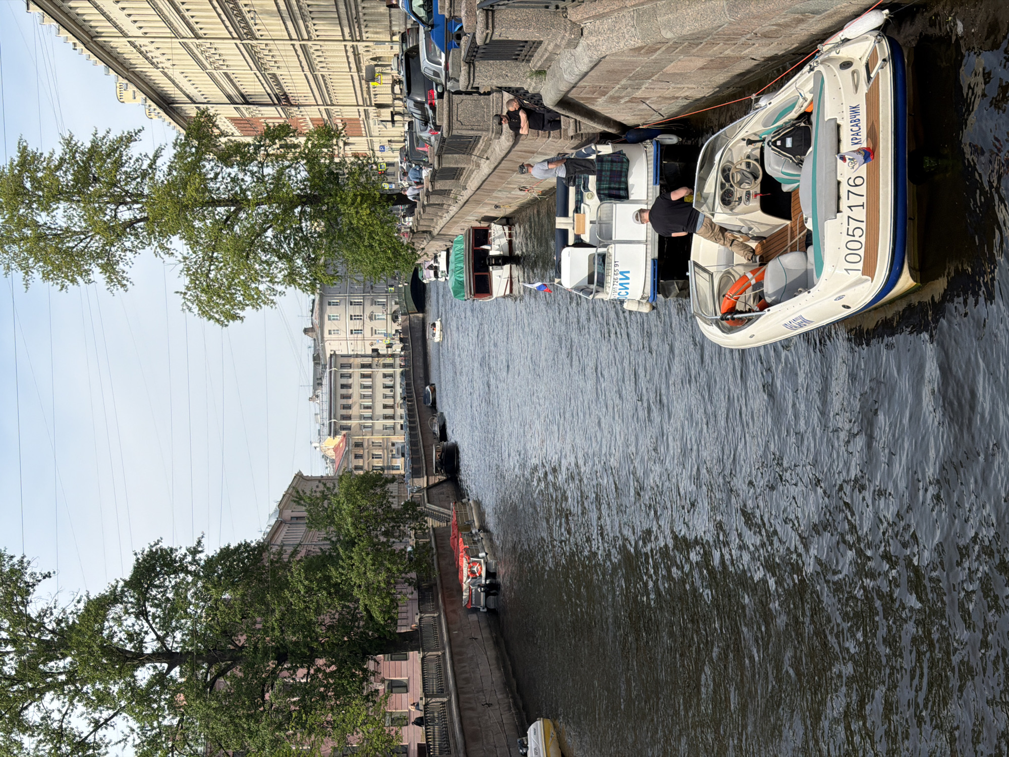 The image captures a view along a canal in Saint Petersburg, Russia, showing boats moored along the embankment. The water is rippled, suggesting a slight breeze or recent boat activity. The embankment is lined with buildings of various architectural styles, indicating a historic urban setting. On the embankment are parked vehicles and pedestrians. Several boats are docked alongside the embankment, with people interacting on or near them. One boat is particularly visible, with the inscription "10057176" on its side. Trees are visible along the embankment, adding greenery to the urban environment. The sky is overcast, casting a soft light over the scene. The general impression is of a typical day along one of Saint Petersburg's many canals, with the mix of historical architecture, waterways, and contemporary activity.