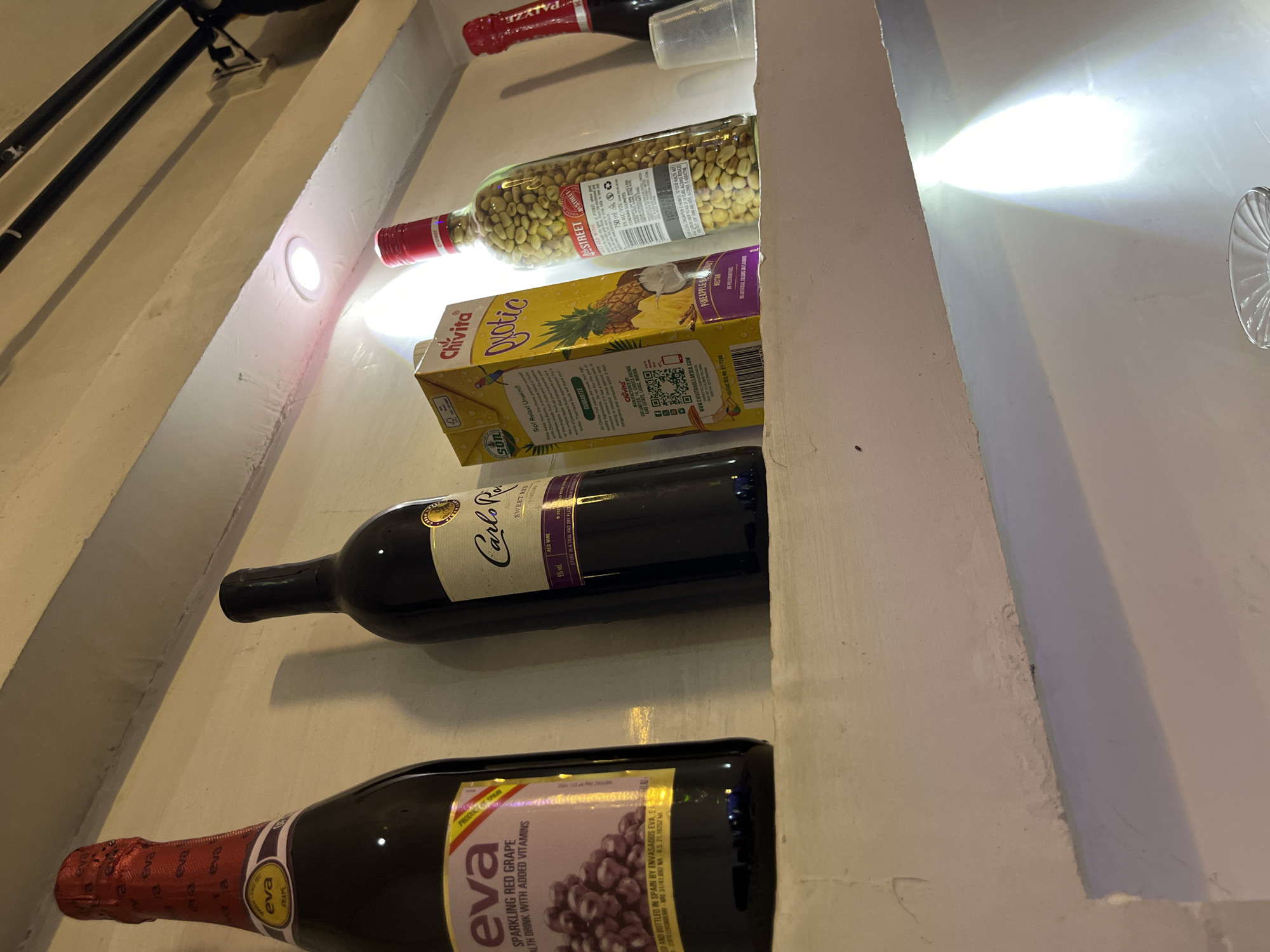 This image depicts a shelf displaying various beverage bottles and a juice box. The scene is indoors, likely a pantry or a display area in a store, given the shelf structure and lighting. The prominent objects include three bottles of wine, one juice box labeled "Chivita Qyotic" with pineapple and coconut imagery, and another bottle containing green olives. The bottles are arranged vertically on the shelf.
The bottle at the bottom is labeled "eva SPARKLING RED GRAPE" and appears to be a wine or sparkling beverage. Above it is a dark bottle of "Carlo Rossi SWEET RED WINE." The bottle containing green olives is a clear glass bottle with a red cap and a label indicating "STREET." The "Chivita Qyotic" juice box is positioned between the olive bottle and a red-capped bottle at the very top of the shelf.
Visible text includes brand names like "eva," "Carlo Rossi," and "Chivita," as well as product descriptions such as "SPARKLING RED GRAPE," "SWEET RED WINE," and "PINEAPPLE NECTAR." There are also indications of origin, such as "PRODUCT OF SPAIN." The lighting suggests it might be daytime or artificial light is being used in a dimly lit area. The overall impression is one of a stocked beverage shelf.
