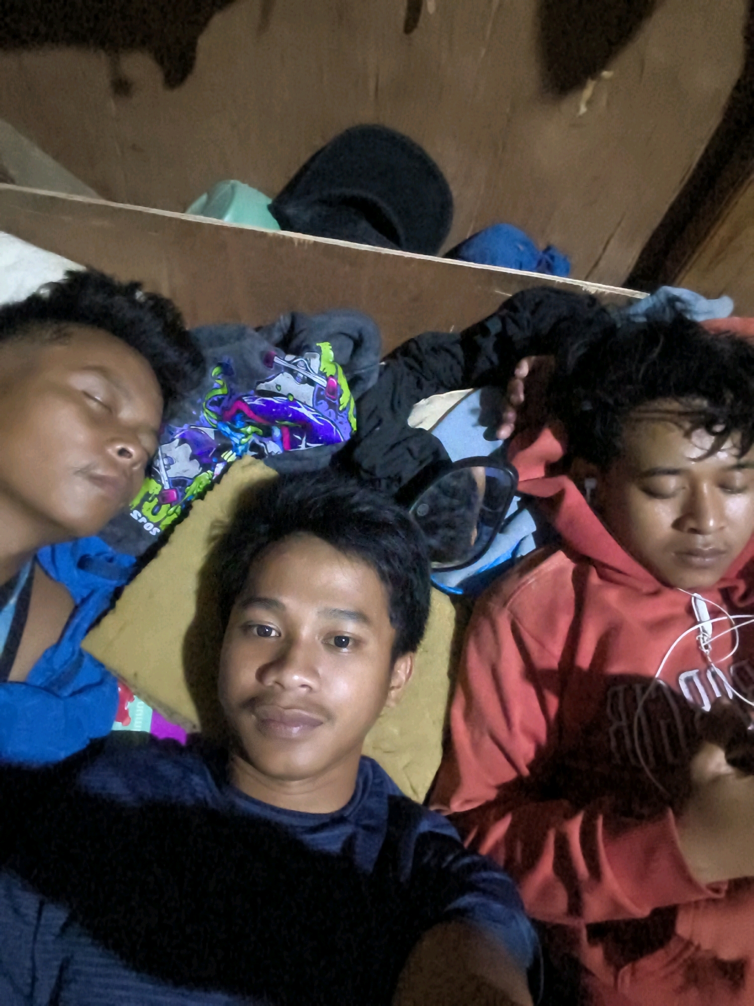 In Bekasi, Indonesia, a selfie captures three young men in what appears to be a cluttered room. The background consists of rough, unfinished wooden shelves stacked with various items, including what seem to be clothing and bags. The lighting is dim, casting shadows that obscure some details of the environment.
The subjects are positioned close together, with one man in the foreground facing the camera, a slight smile on his face. His dark t-shirt is visible. To his left, another man is lying down with his eyes closed, appearing to be asleep. He is wearing a blue shirt. To the right of the foreground subject, a third man is also lying down, his eyes closed, and wearing a red hoodie. White earbuds are visible in his ears. Scattered around the men are articles of clothing, including a gray hoodie with a colorful graphic design. A pair of dark sunglasses is also visible on a piece of fabric. The overall impression is one of casualness and informality, possibly in a dorm room or a similar living space.