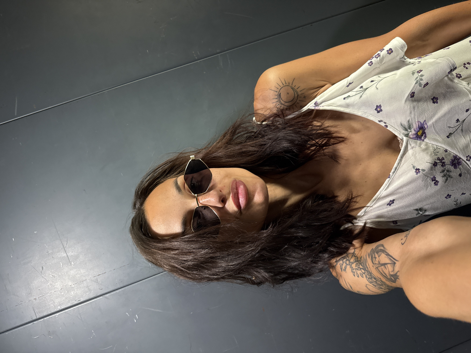 A woman with long, dark, wavy hair, wearing sunglasses and a white floral dress, poses for a selfie in front of a dark grey, corrugated metal wall. She has tattoos on her left arm and right shoulder. The image was taken in Pleven, Bulgaria.
