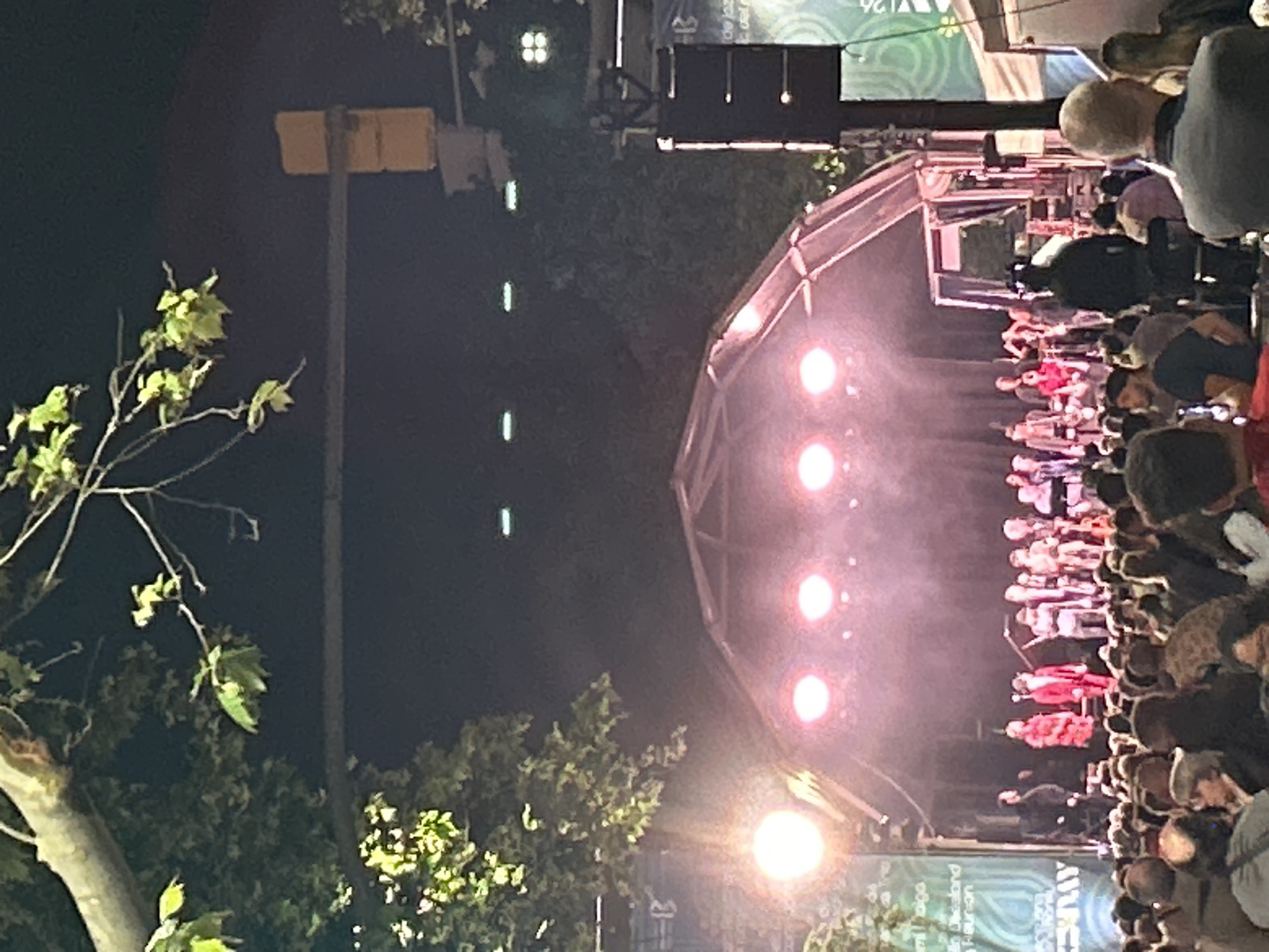 The image depicts an outdoor concert at night. A large crowd faces a brightly lit stage with a domed structure supporting lighting fixtures. The crowd is dense, with people standing close together. The stage has several figures likely performing. A few trees are visible in the foreground and to the left. In the background, there's a street lamp and other light sources. While specific clues from the image are limited, based on the event banners at the top of the stage, it's likely the image was taken in Barcelona, Spain.