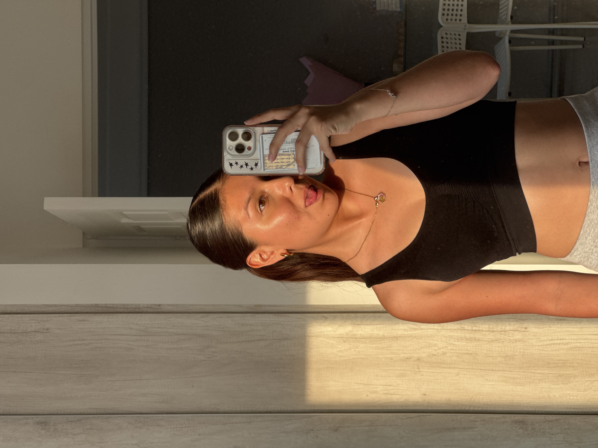 A woman is taking a selfie with a phone. She is wearing a black sports bra and grey sweatpants. The photo appears to be taken indoors, next to a wooden surface with strong sunlight casting shadows. A partial view of a window or a door is seen in the background. The scene doesn't provide enough context to locate it in Ocotlán.