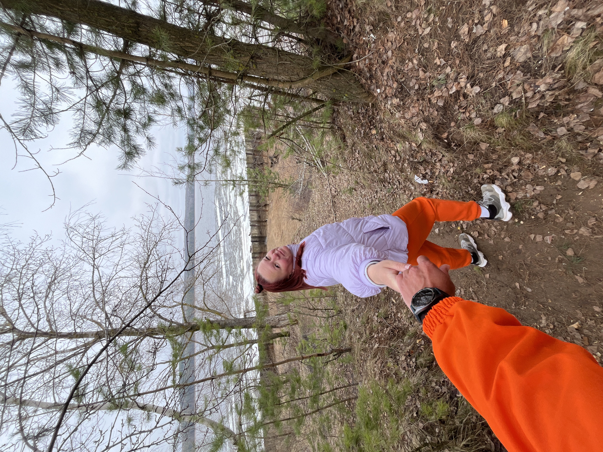 The image, rotated 90 degrees clockwise, captures a first-person perspective of two individuals holding hands in a natural, outdoor setting, likely in Chaikovsky, Russia. The person holding the camera is only visible by their right arm, clad in a vibrant orange jacket and sporting a black wristwatch. They are holding the hand of a woman who stands before them, facing the camera.
The woman has reddish-brown hair styled into two buns and is dressed in a light purple quilted jacket, bright orange pants, and grey and white chunky sneakers. Her expression is calm, with a slight smile, as she looks directly towards the viewer.
The background reveals a wooded area with a mix of pine trees and bare deciduous trees, indicating a late autumn or early spring season. The ground is covered with dry, fallen leaves and sparse, dry grass. Beyond the trees, a large body of water, partially covered with chunks of ice, stretches towards a grey, overcast sky. The overall lighting suggests it is daytime, under a cloudy sky, contributing to a cool, possibly chilly, atmosphere. The act of holding hands conveys a sense of connection and companionship between the two people during their outdoor excursion. There is no visible text in the image.