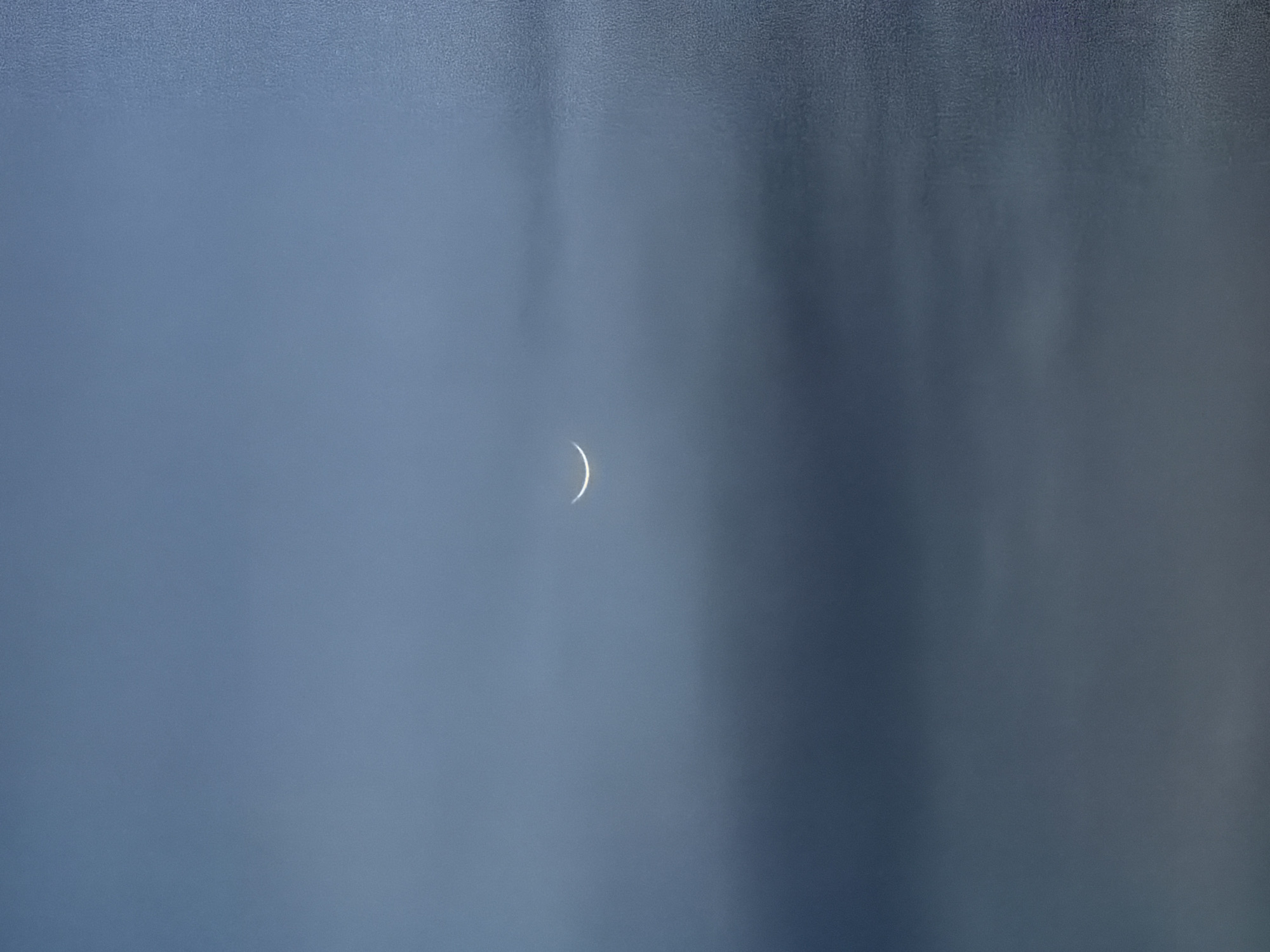 A crescent moon is visible in a hazy, overcast sky over Baan Bang Rak, Thailand. The moon is a thin sliver of light against the muted blue-gray of the clouds. The overall impression is one of a soft, diffused light with a sense of stillness. There are no people or man-made objects clearly discernible in the frame.