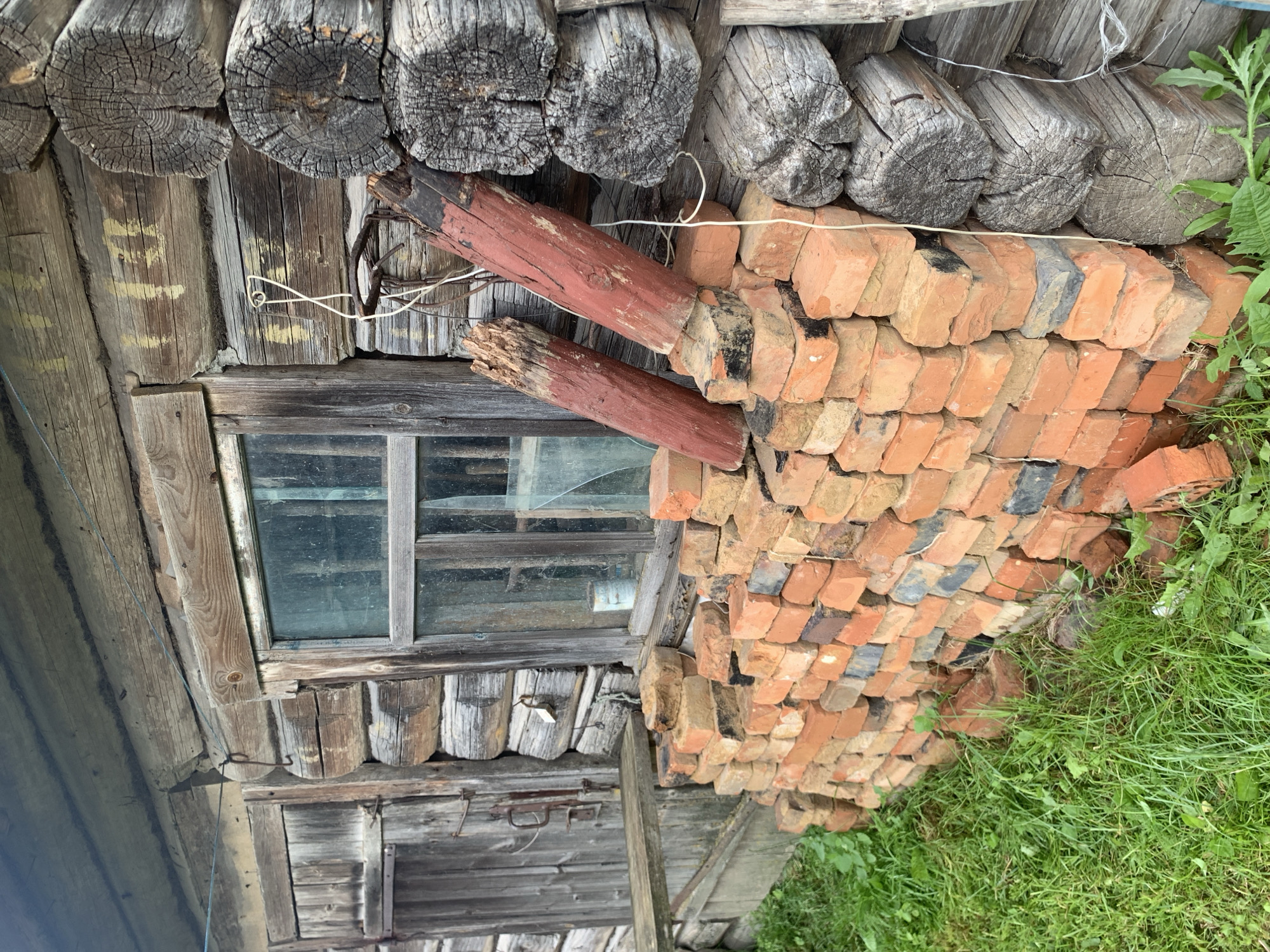 The image depicts a section of an old, weathered log cabin, likely in a rural setting within Vidzaŭski sel'ski sovet, Belarus. The cabin's walls are constructed from large, rough-hewn logs, showing signs of age and exposure to the elements. A window with wooden panes and dusty glass is visible on the wall. In front of the cabin, a large stack of reddish-orange bricks is piled haphazardly against the foundation. Two weathered wooden posts, painted red at their lower ends, lean against the stacked bricks and the cabin wall. The ground is covered in lush green grass, with some weeds and clover growing amongst the bricks. The overall impression is one of rural decay and rustic simplicity. There are no people or animals visible in the image.