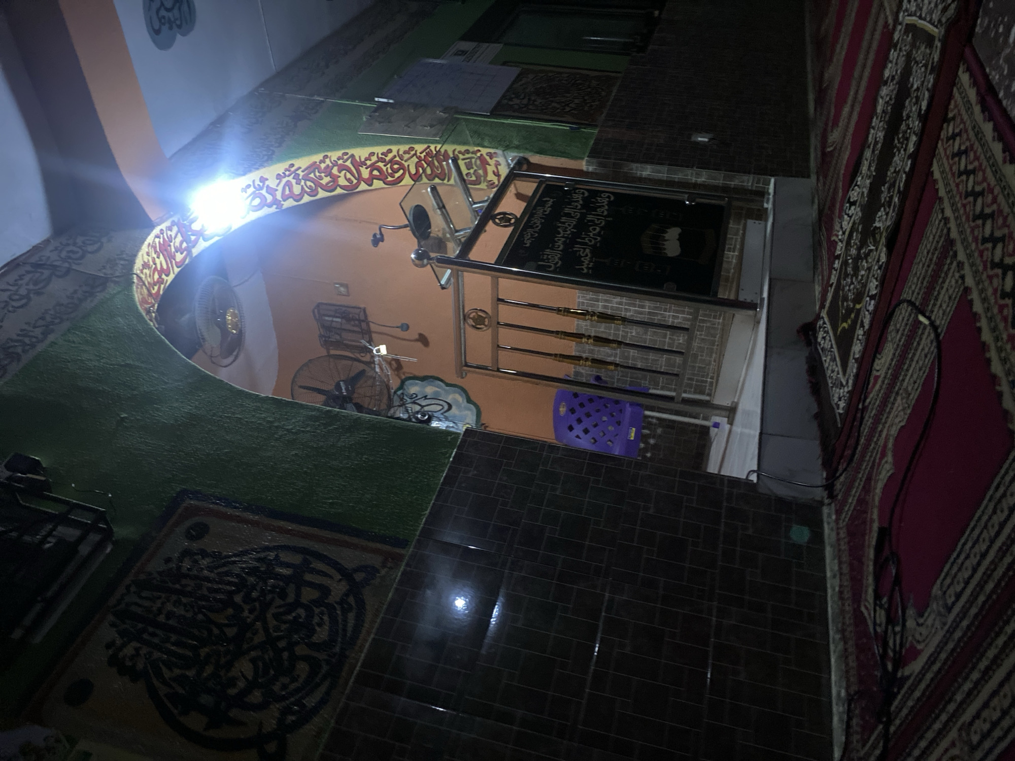 The photograph shows the interior of a building, likely a mosque, in Lagos, Nigeria. The walls are painted with intricate designs and religious inscriptions in Arabic script. A large arched structure with decorative elements forms a prominent feature, bathed in bright light. There is a ceiling fan and metal grilles in the background. The floor is tiled with dark, rectangular tiles in the foreground and also includes decorative rugs and carpets in shades of red, black, and beige. There is a blue plastic chair and a metal railing with a dark signage attached. The overall lighting is dim, except for the brightly illuminated archway.
