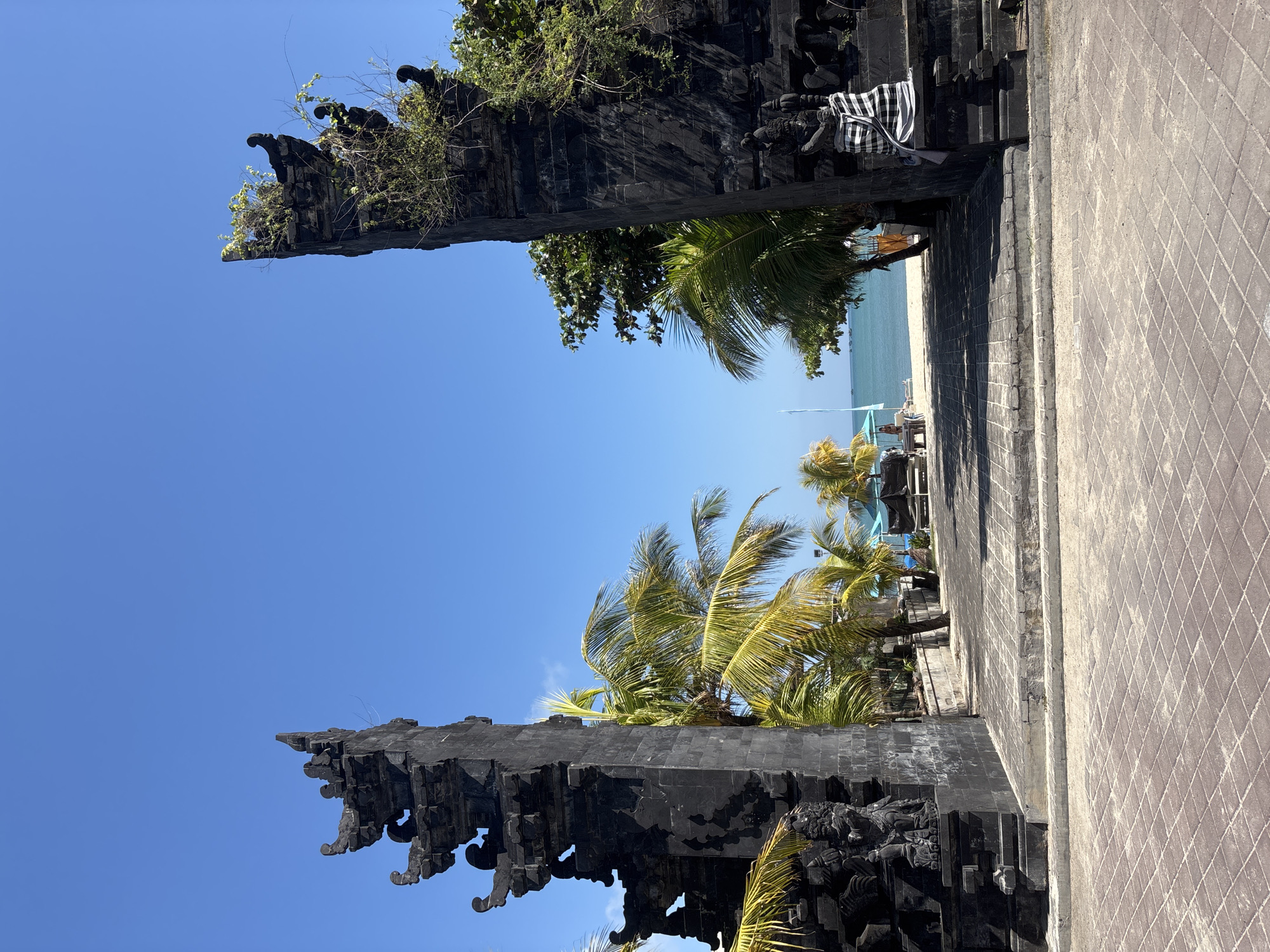 The image shows a traditional Balinese temple gateway, known as Candi Bentar, located in Kuta, Indonesia. The gateway is constructed from dark volcanic stone and is adorned with intricate carvings and statues. The gateway opens to reveal a view of a sandy beach with palm trees, the turquoise ocean, and a clear blue sky. In the distance, several boats are visible on the water. There are a few people on the beach, some sitting on lounge chairs and others walking. The foreground is paved with stone tiles. The overall scene conveys a serene and tropical atmosphere.