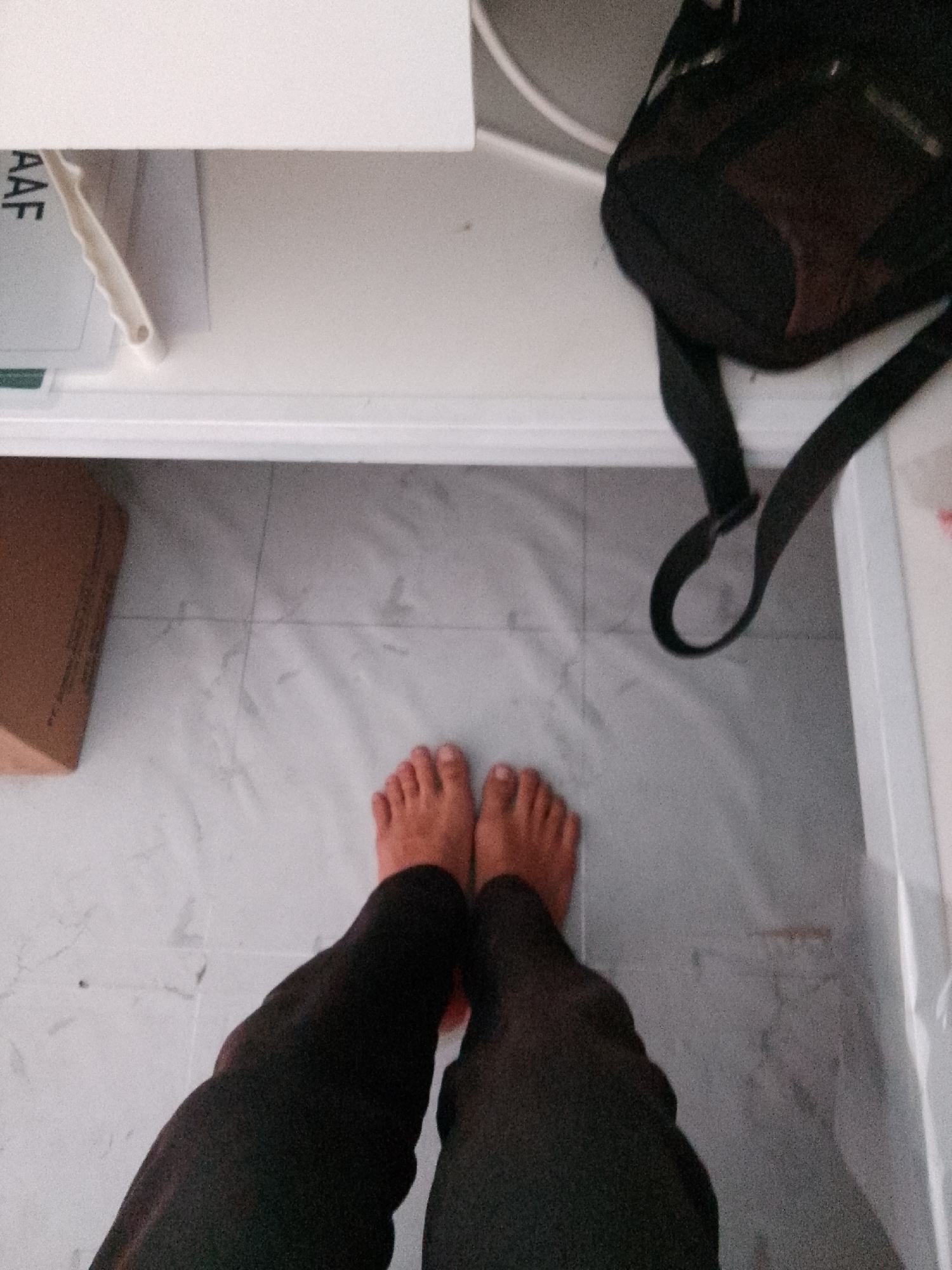 An overhead shot shows a person's bare feet and lower legs standing on a marbled tile floor. The person is wearing dark pants. To the left of the feet, a brown cardboard box sits on the floor. In the background, a white shelf is visible with some papers on it, and a black backpack with straps hangs from it. The scene appears to be indoors, possibly in a storage area or a room with limited space. The lighting is dim, casting shadows on the floor. The image does not provide any specific details about the location in the city of Trenggalek, Indonesia.