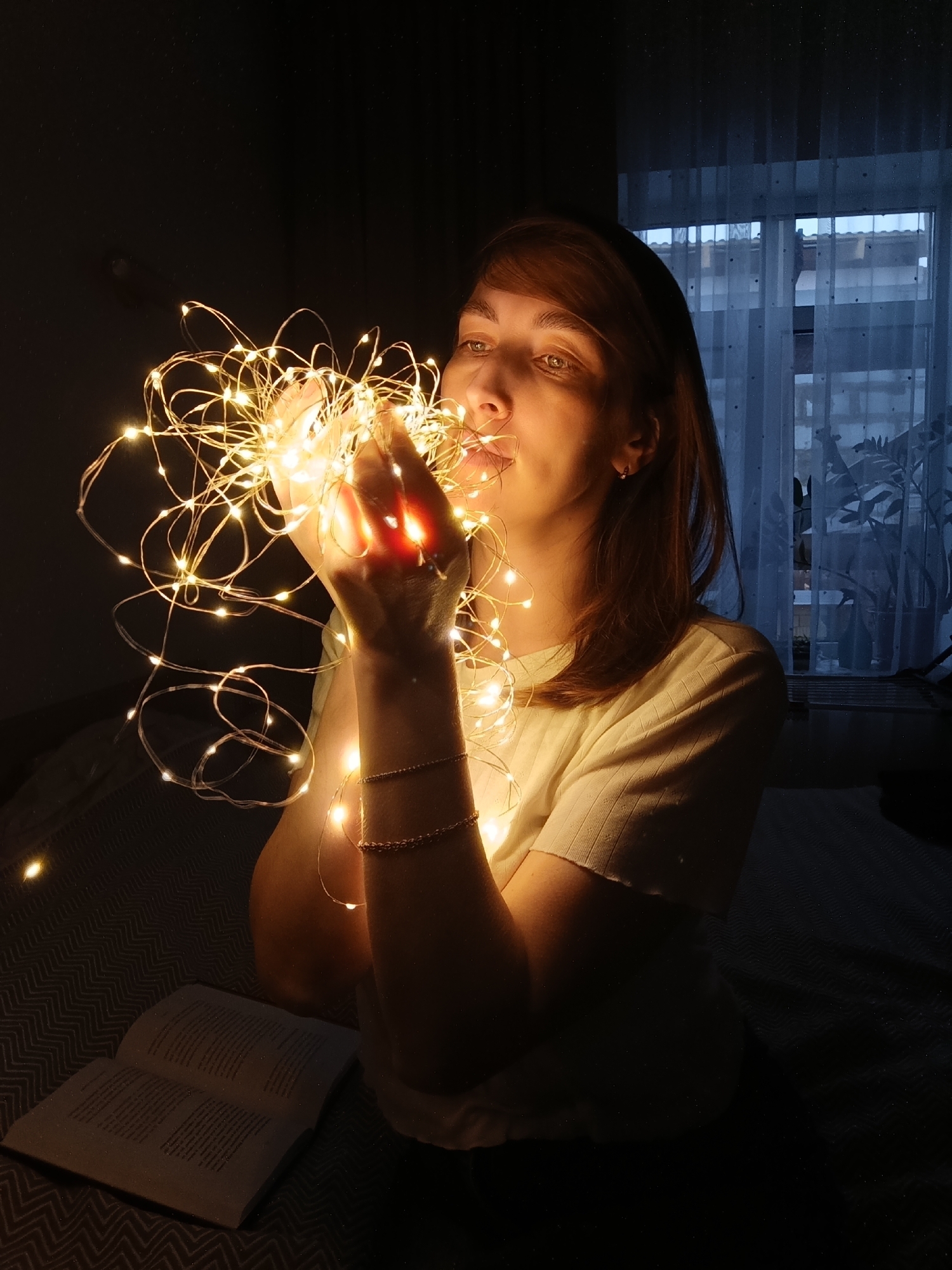 A woman with reddish-brown hair and light skin holds a tangle of warm white LED string lights in both hands, close to her face, in a dark interior. She wears a light-colored, short-sleeved shirt and a gold bracelet on each wrist. An open book lies on a chevron-patterned surface, possibly a bedspread, to the lower left of the frame. In the background, a window with a sheer white curtain is visible, suggesting it is daytime outside. A dark shape resembling a potted plant stands near the window. There is a light fixture visible at the top left edge of the image. The location is likely in an indoor setting. The city of Chaikovsky, Russia, is not evident within the image.