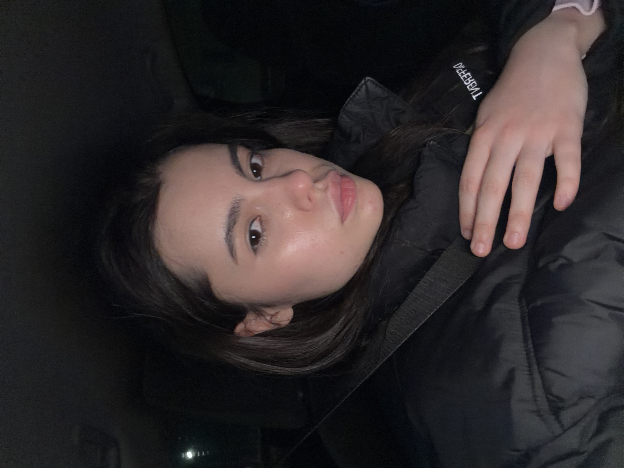 A young woman is captured in a close-up, self-portrait style image, seemingly lying down or leaning back in a dark environment, possibly inside a vehicle at night in Chaikovsky, Russia. She looks directly at the camera with dark eyes and a soft, slightly pouting expression. Her dark hair frames her face, and her complexion is fair.
She is wearing a black, puffy winter jacket or coat, indicative of cold weather. A dark strap, resembling a seatbelt or bag strap, crosses her body. Her right hand rests on her jacket, revealing a light-colored, possibly pink or white, cuff peeking from beneath her sleeve. On the jacket's chest area, white text is visible, appearing mirrored but readable as "DIFFERENT."
The background is very dark, suggesting minimal lighting, with only a few faint, blurry glints of light in the distance that could be streetlights or other reflections. The overall impression is an intimate, calm moment captured in low light.