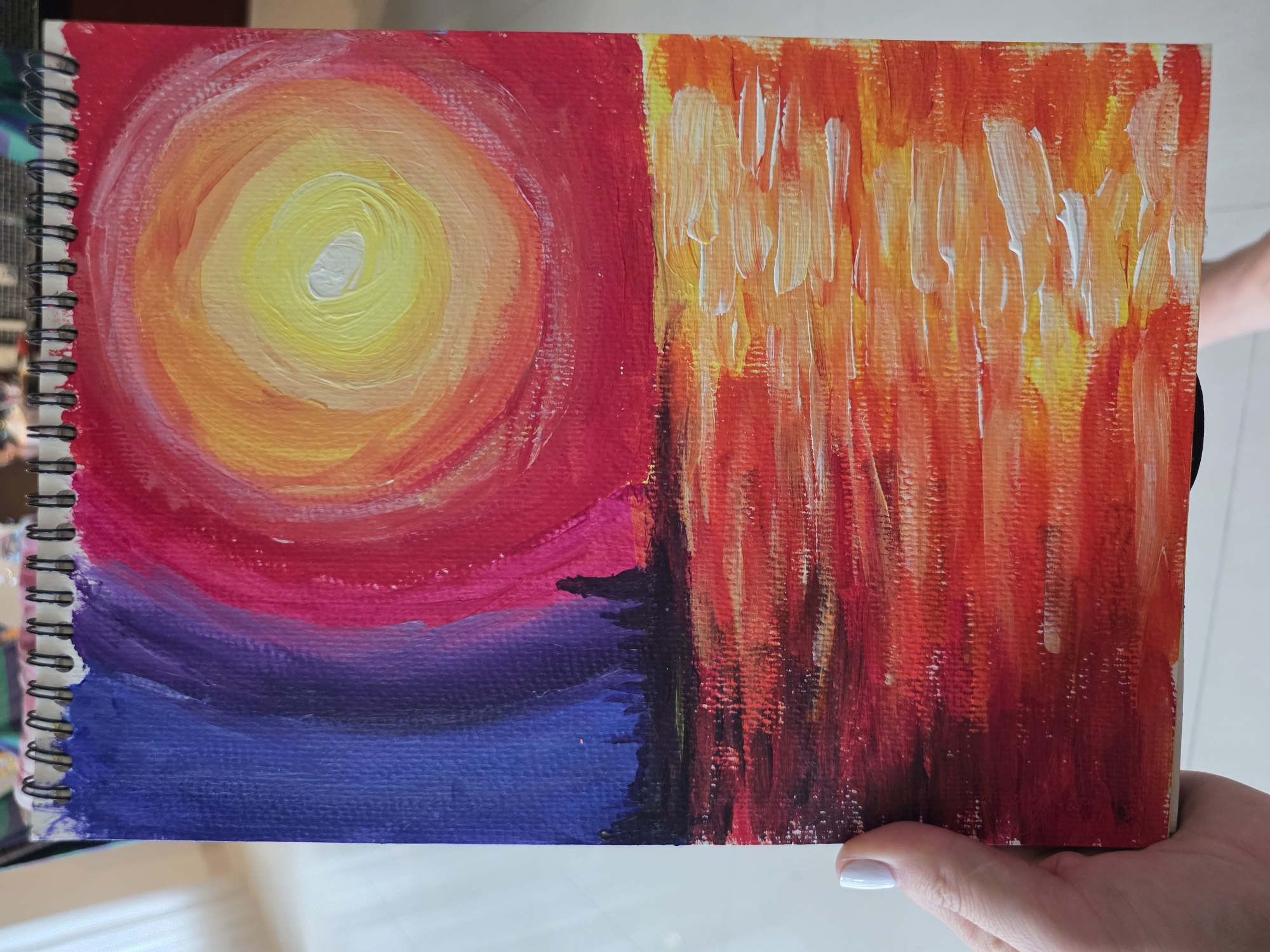 A photograph shows an open sketchbook displaying a painting. The painting is divided into two distinct sections. On the left is a circular image of a sunset. The center of the circle is a bright yellow, surrounded by rings of orange and then red. Below this are bands of pink, purple, and blue. The right side of the painting features vertical strokes of paint in shades of red, orange, yellow, and white, creating a textured effect. The background is out of focus, but appears to be an indoor space. The sketchbook is held by a hand with light skin and a white fingernail. The image was not taken in North Kuta, Indonesia.