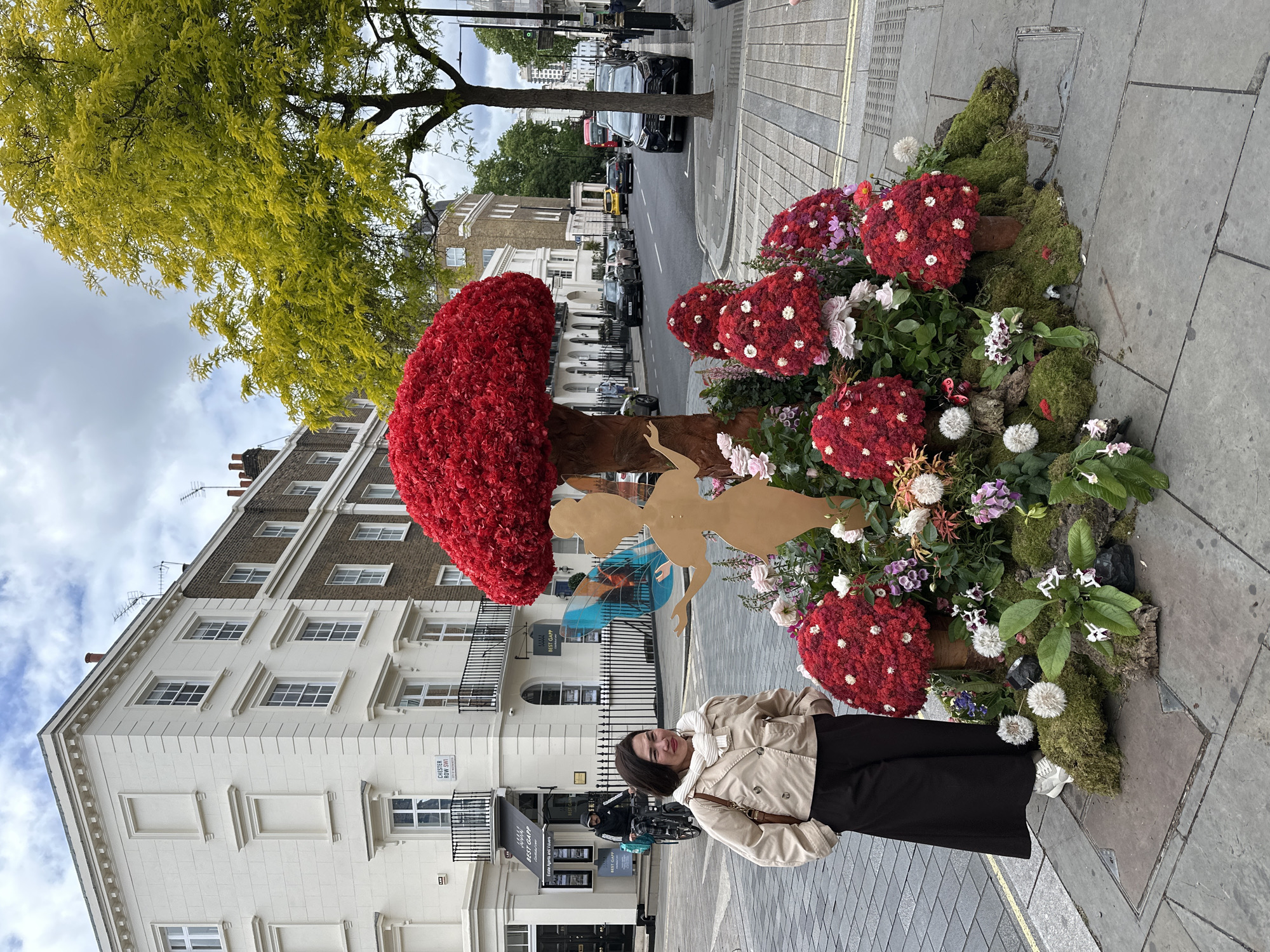 The scene is an outdoor setting in London, United Kingdom. A woman stands on a sidewalk next to a large, floral art installation resembling a tree and several toadstools. The tree has a brown trunk and a large, red flower-covered head. The toadstools are also covered in red flowers with white accents. The installation is surrounded by other flowers, plants, and moss.
Behind the woman and the installation is a row of multi-story buildings, some with white facades and black iron balconies. The street is visible between the buildings, with cars parked along the sides. A tree with yellow-green leaves is also visible above the buildings.
The woman is wearing a beige jacket, a white scarf, and a long black skirt. She is smiling and looking at the camera. The sign near the door of a building behind reads "BEST GAPP Estate Agents and Valuers". Another sign reads "Chester Row SW1W".