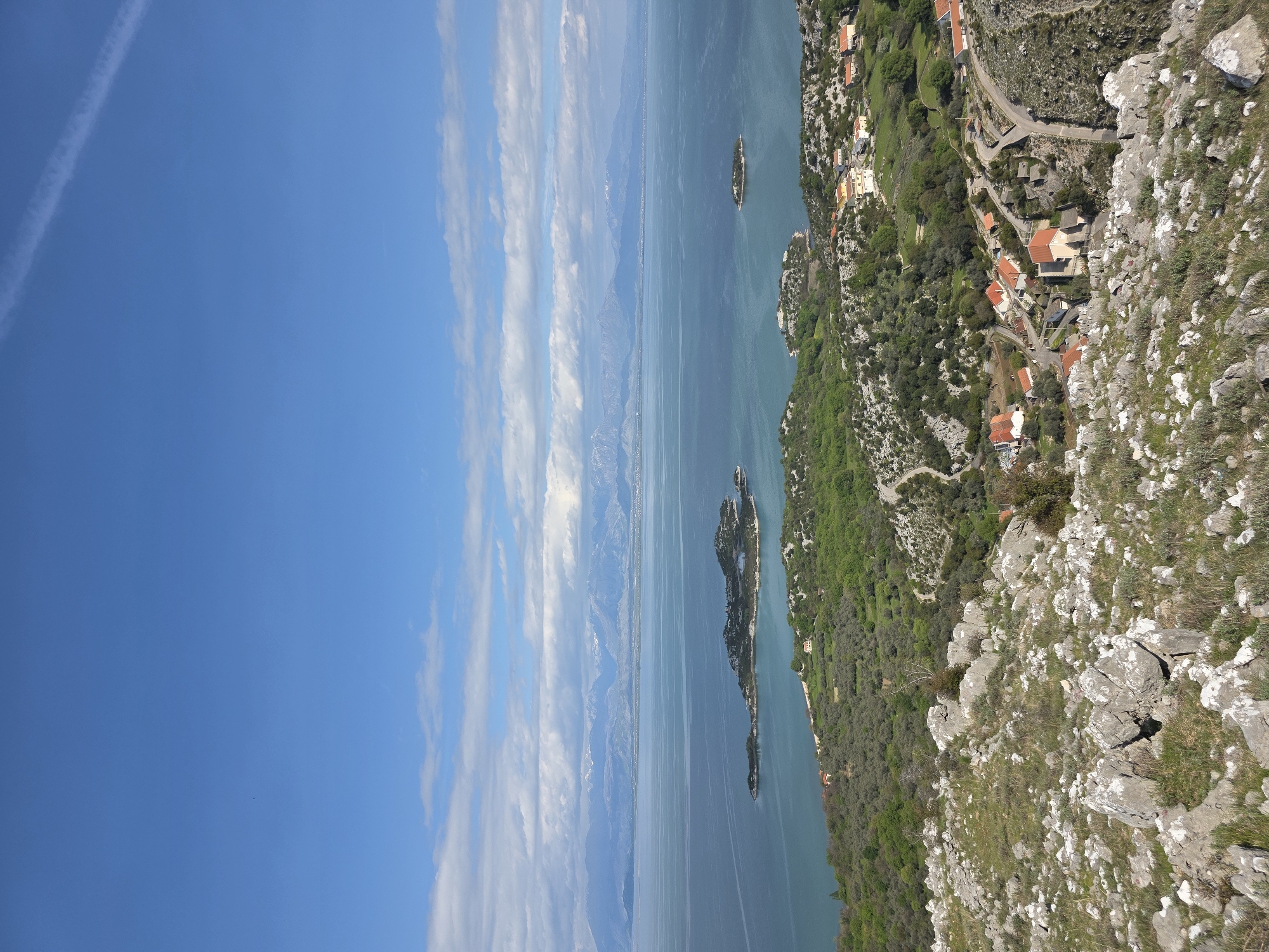 The image depicts a view from a high vantage point in Donji Murići, Montenegro, showcasing a coastal landscape. The sky is clear blue with some streaky clouds. Below, there's a body of water that appears to be a bay or part of a larger lake or sea, with islands dotting the surface. The land is rugged, with rocky terrain and patches of green vegetation. There are clusters of buildings with red-tiled roofs nestled among the trees. A winding road connects the buildings. The overall impression is that of a rural coastal area with a mix of natural and human elements.
