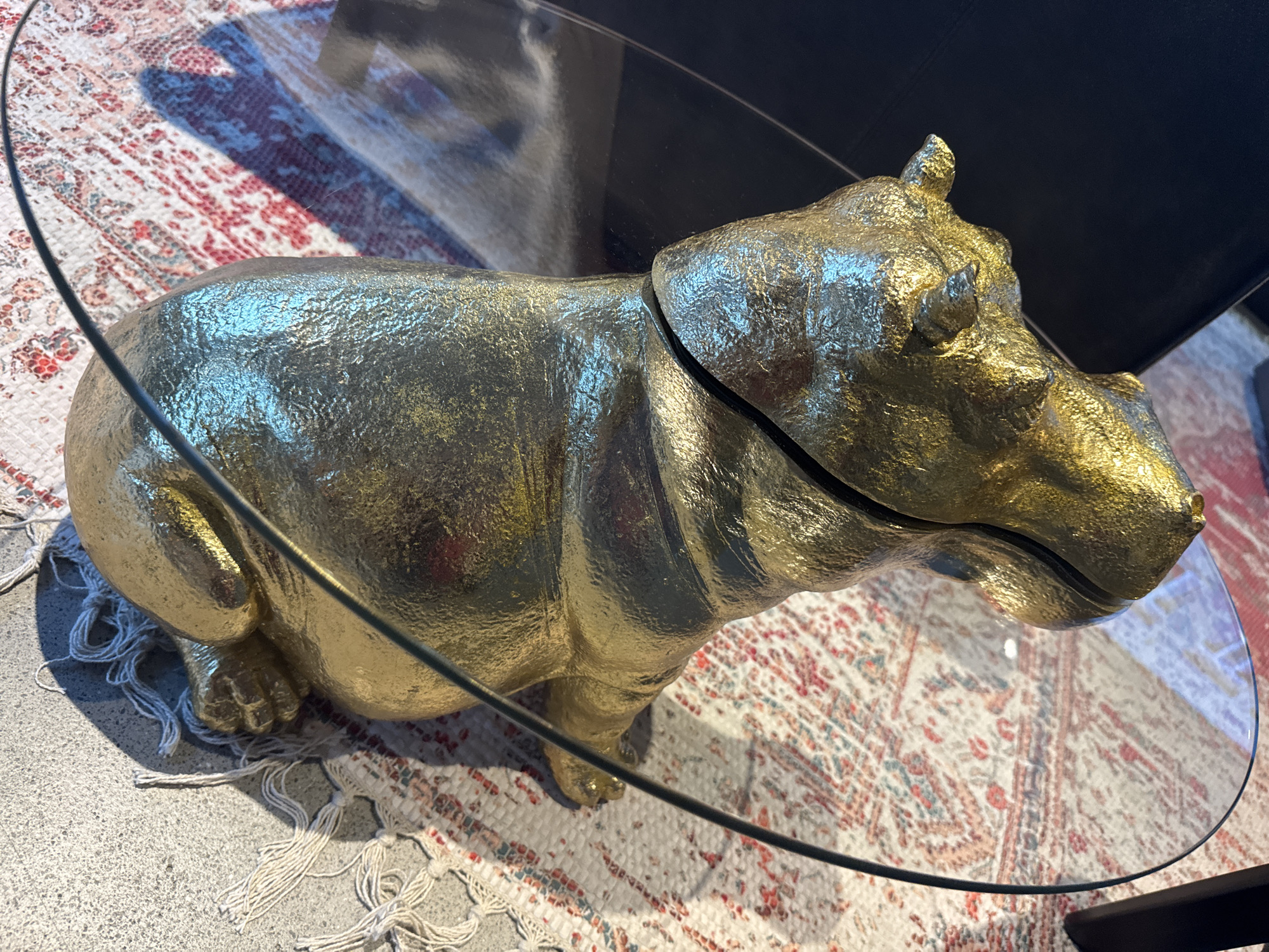 A close-up, top-down view of a golden, segmented hippopotamus sculpture is shown. The sculpture is placed on a patterned rug, which in turn is on a rough, textured surface, possibly concrete. A thin, clear glass tabletop partially covers the sculpture and the rug, creating reflections. The sculpture itself appears to have a rough, metallic texture and is split into two main parts. One part, the head, rests on the rug, while the other, the body, is partially obscured by the glass. The lighting suggests an indoor setting with strong shadows cast by the sculpture and the glass. No people are visible in the image, and the scene appears to be a still-life arrangement of decorative objects. The provided context places this scene in the city of Nendeln, Liechtenstein.