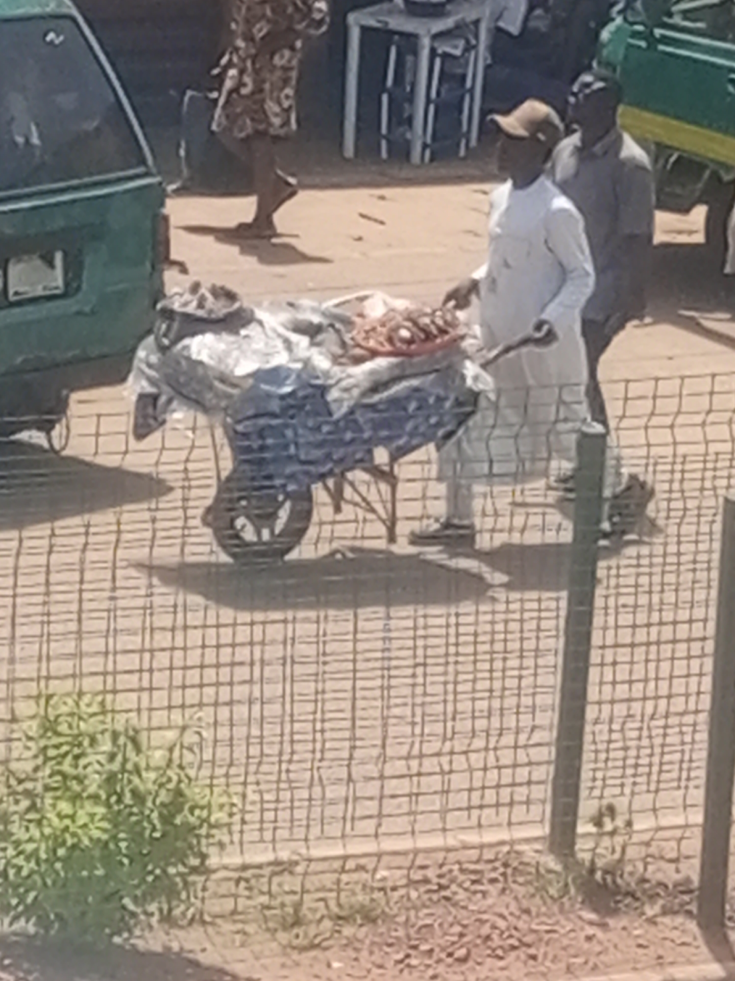 A person wearing a white robe and a brown cap stands near a wheelbarrow laden with goods wrapped in plastic. The wheelbarrow has a large wheel with spokes visible. To the right of this person, another person wearing a gray shirt stands. In the background, a green vehicle is partially visible on the left, and another green vehicle with a yellow trim is on the right. A person in a patterned dress walks in the background. The scene appears to be an outdoor market setting, possibly in Bukuru, Nigeria, given the context. The ground is dusty, and a wire fence is in the foreground, obscuring some of the view. The lighting suggests it is daytime.