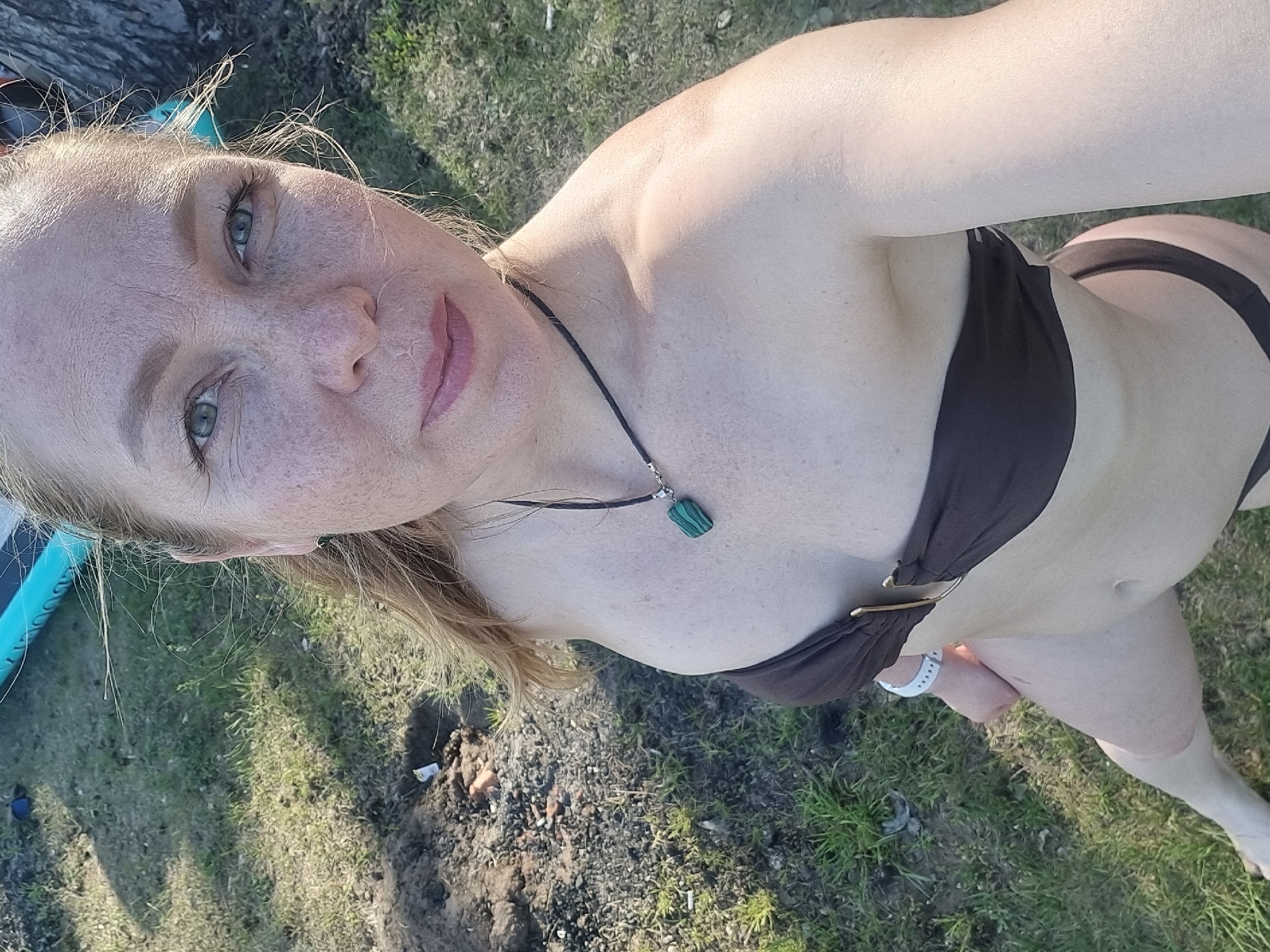 A fair-skinned woman with freckles poses in a brown bikini. She has light blonde hair and green eyes, and is wearing a necklace with a green pendant and a white smartwatch. The background shows a grassy area and possibly the edge of a body of water. The location is Chaikovsky, Russia.