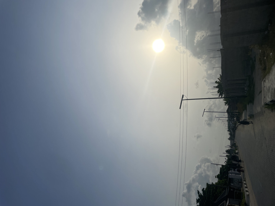 The image depicts a street scene in Mosafejaw, Nigeria, under a partly cloudy sky with the sun prominently visible in the upper right quadrant. The sky transitions from a light blue-grey at the top to a hazy yellow-white near the bright, circular sun, which emits a distinct lens flare. Scattered cumulus clouds, ranging from white to dark grey, are present across the mid-ground of the sky.
A paved road, constructed from interlocked pavers, extends from the foreground into the distance, exhibiting a slight curve. Overhead power lines span horizontally across the mid-ground. Along the left side of the road, a series of single-story structures are partially visible behind green foliage.
On the right side of the paved road, a dirt path runs parallel. Multiple dark-colored streetlights, each equipped with an integrated solar panel, stand vertically along this path, receding into the background. Adjacent to the closest streetlights, a partially constructed building with exposed rebar is visible.
Several individuals are present on the road, silhouetted against the bright lighting. One person, dressed in dark attire, walks away from the viewer on the right side of the paved road. Additional human figures are discernible walking further in the distance. The scene is characterized by strong backlighting from the low sun, creating significant contrast.