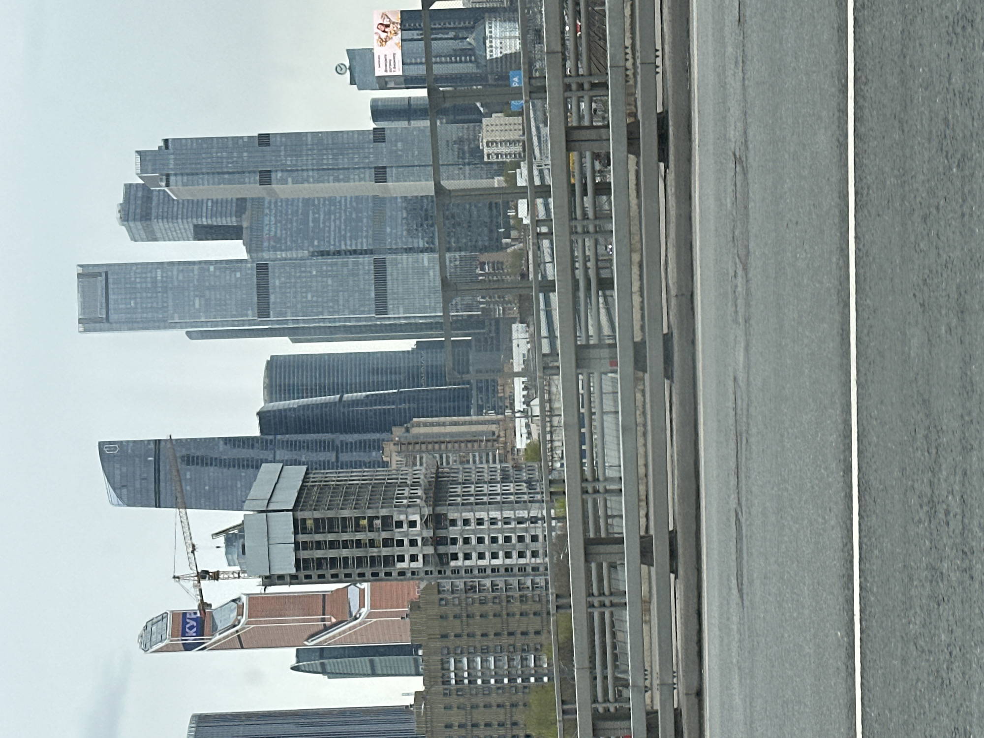 The image shows a view of the Moscow International Business Center (Moscow City) from a bridge or elevated roadway. The foreground consists of a gray concrete road with a white lane marker and a metal guardrail. In the background, several skyscrapers of varying heights and architectural styles are visible, including some buildings under construction with a yellow tower crane. A billboard is visible on the top of one of the buildings. The sky is overcast and gray.