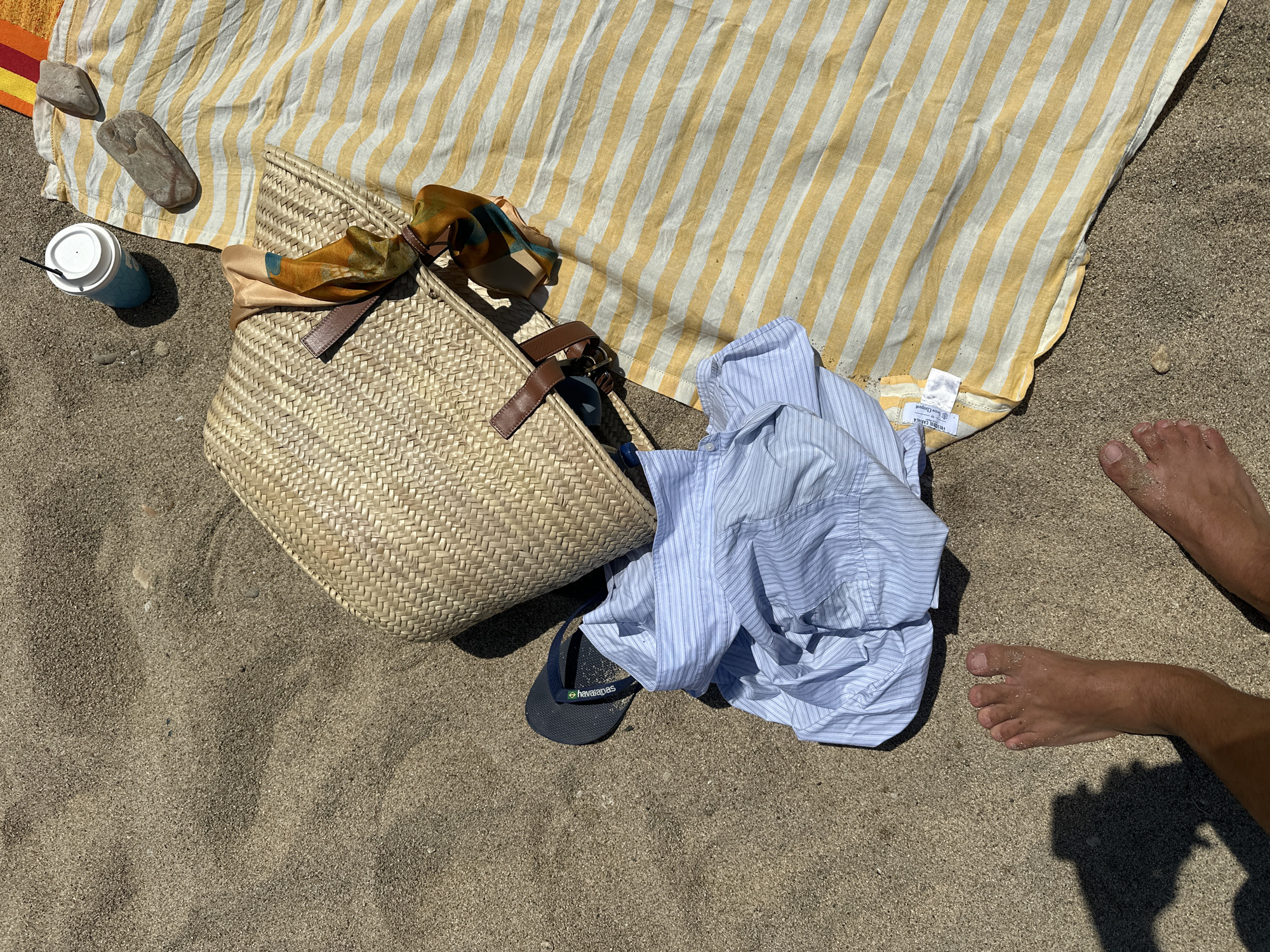 The image depicts a beach scene in Ano Mera, Greece. A woven straw bag with brown leather straps sits on a yellow and white striped beach towel. A blue cup with a white lid and a straw, two grey stones, a blue and white striped shirt, and a pair of dark flip-flops are also on the towel. A person's feet, with visible sand on them, are in the lower left and right of the frame, casting shadows on the sand. The environment is sandy with some small pebbles.