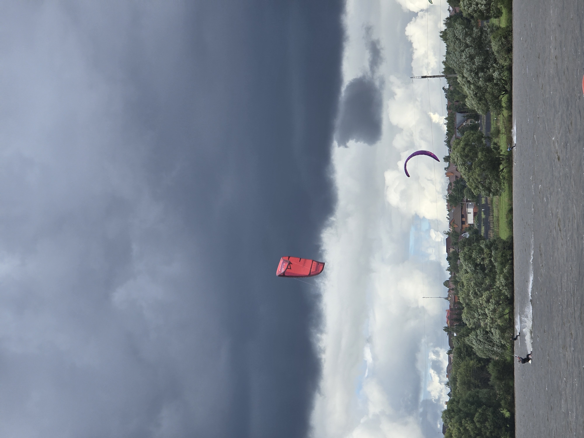 The image depicts a body of water with several individuals kitesurfing. In the foreground, a kitesurfer is actively engaged in the sport, creating a spray of water behind them. Further in the distance, another kitesurfer can be seen, with a purple kite visible in the sky. A red kite is also prominent in the upper portion of the image. The background features a shoreline lined with trees and houses, indicative of a settlement. The sky is overcast with dark storm clouds dominating the upper half of the frame, while lighter clouds are present lower down. The location is identified as Novoseltsevo, Russia.
