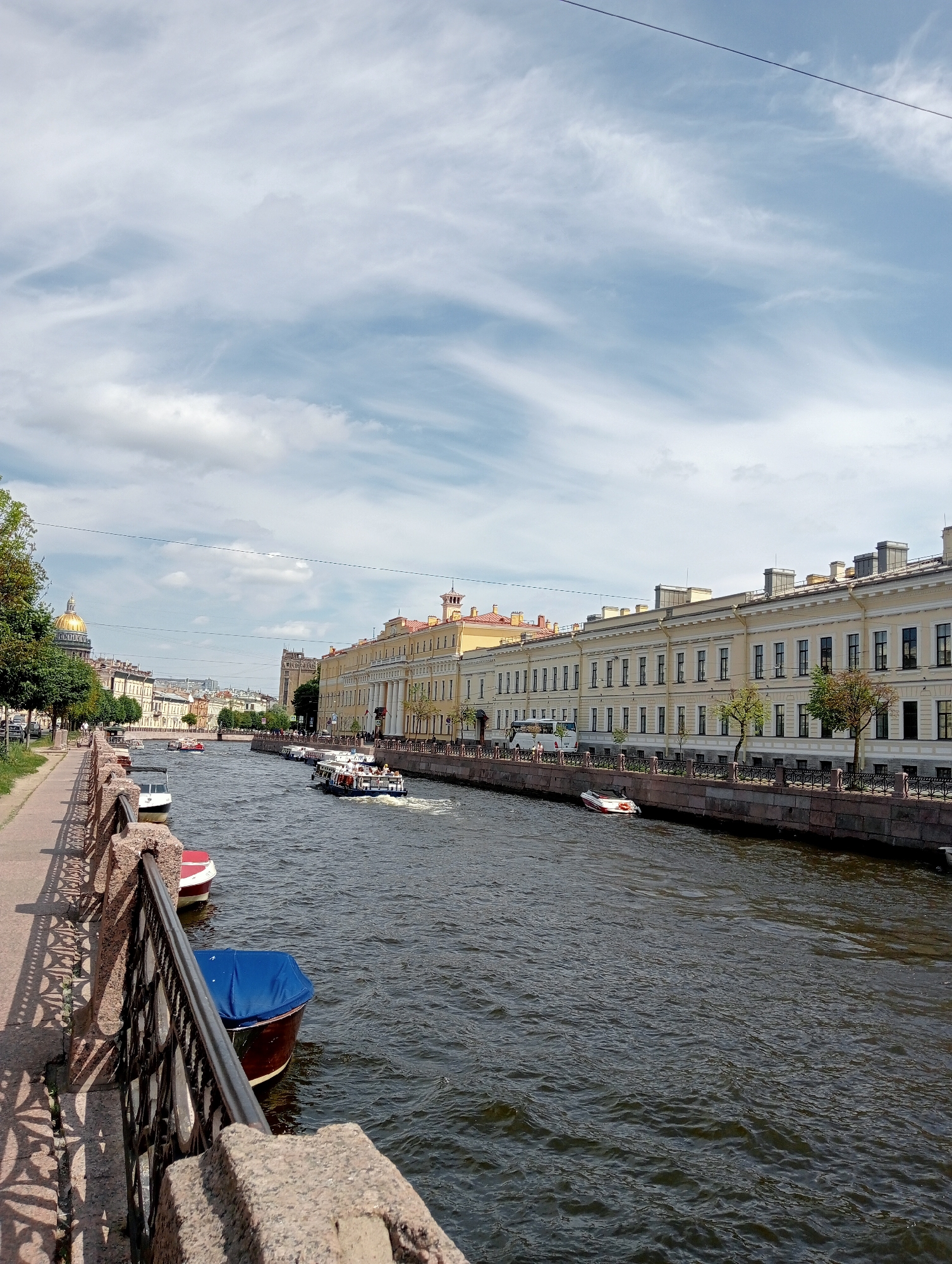 The image captures a view of the Griboyedov Canal in Saint Petersburg, Russia, on a partly cloudy day. A promenade lines the left bank of the canal, featuring a stone embankment with an ornate metal railing. Trees with green foliage grow along the promenade. On the right bank, a long, pale yellow building with numerous windows and a classical facade stretches into the distance, its architecture characteristic of Saint Petersburg.
Several tour boats are navigating the canal, some carrying passengers. In the foreground on the left, a dark-colored boat is partially covered with a blue tarp. Further down the canal, a white boat with red accents is docked. In the middle distance, a larger white tour boat with passengers moves down the canal.
In the background, the iconic golden dome of Saint Isaac's Cathedral is visible on the left, indicating the general location within the city. The sky is a mix of blue and white clouds. The water in the canal is dark and choppy, reflecting the sky and surrounding architecture. The scene depicts a typical day of tourism and urban life along one of Saint Petersburg's historic waterways.