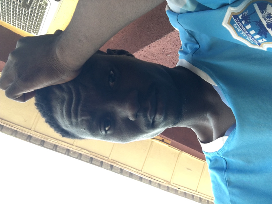 A young adult male with dark skin is depicted in a slightly upward-angled, close-up shot. He has short, dark, curly hair and prominent wrinkles across his forehead. His right hand, with fingers curled, rests on the top of his head. He wears a light blue short-sleeved shirt with a white collar. An emblem on his right sleeve prominently displays "Bahir Dar Kenema F.C." and other less legible text, alongside a graphic of a cityscape or waterfront. The subject maintains a neutral expression while looking directly forward. The outdoor environment includes a bright, overexposed sky in the upper left background, beneath which a building structure with light yellowish-brown horizontal paneling is visible. To the upper right, a reddish-brown textured wall appears, partially obscuring a white, slatted object, possibly a vent. Strong overhead lighting casts shadows across the subject's face, particularly on his right side. This scene is located in Pindiga, Nigeria.